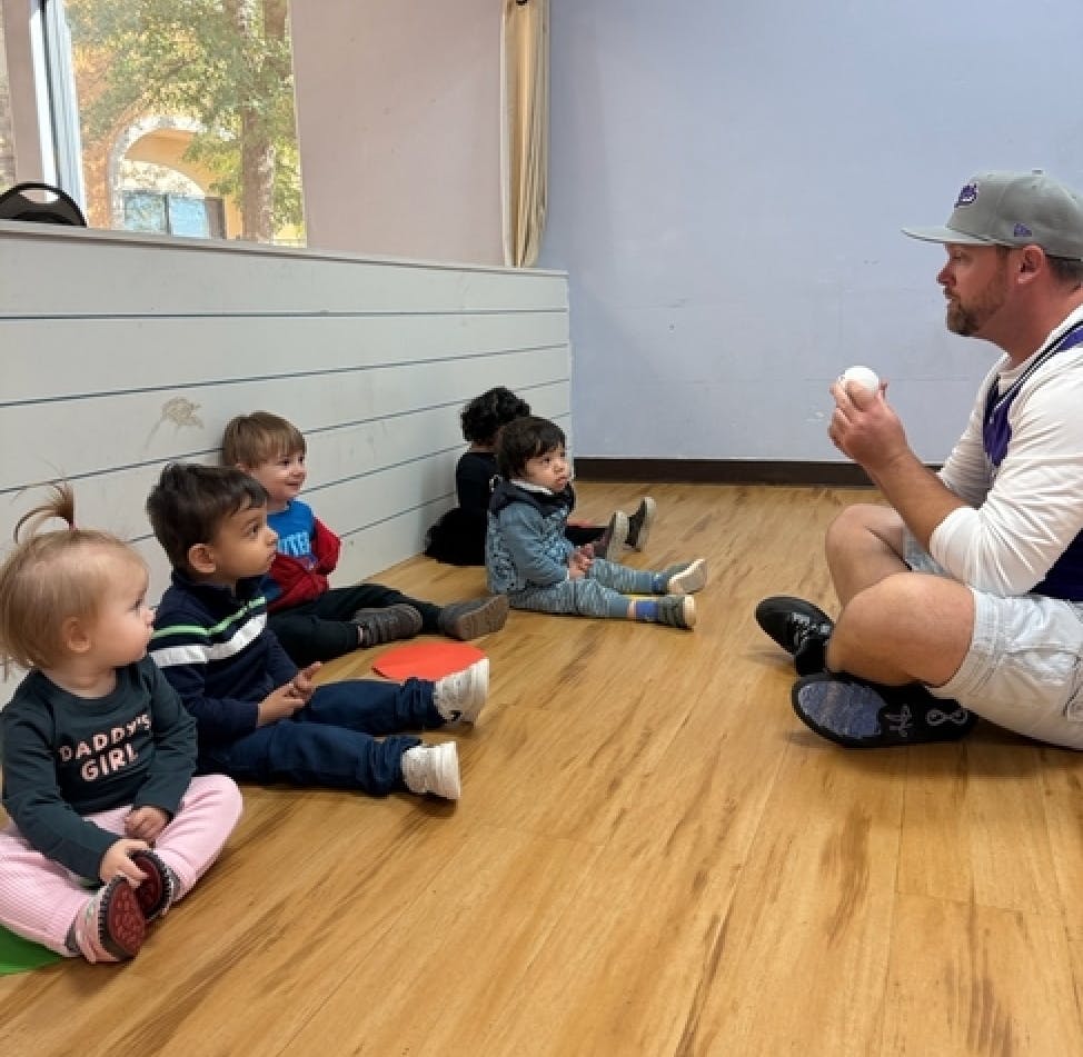 Adult holding a baseball talking to children sitting on the floor
