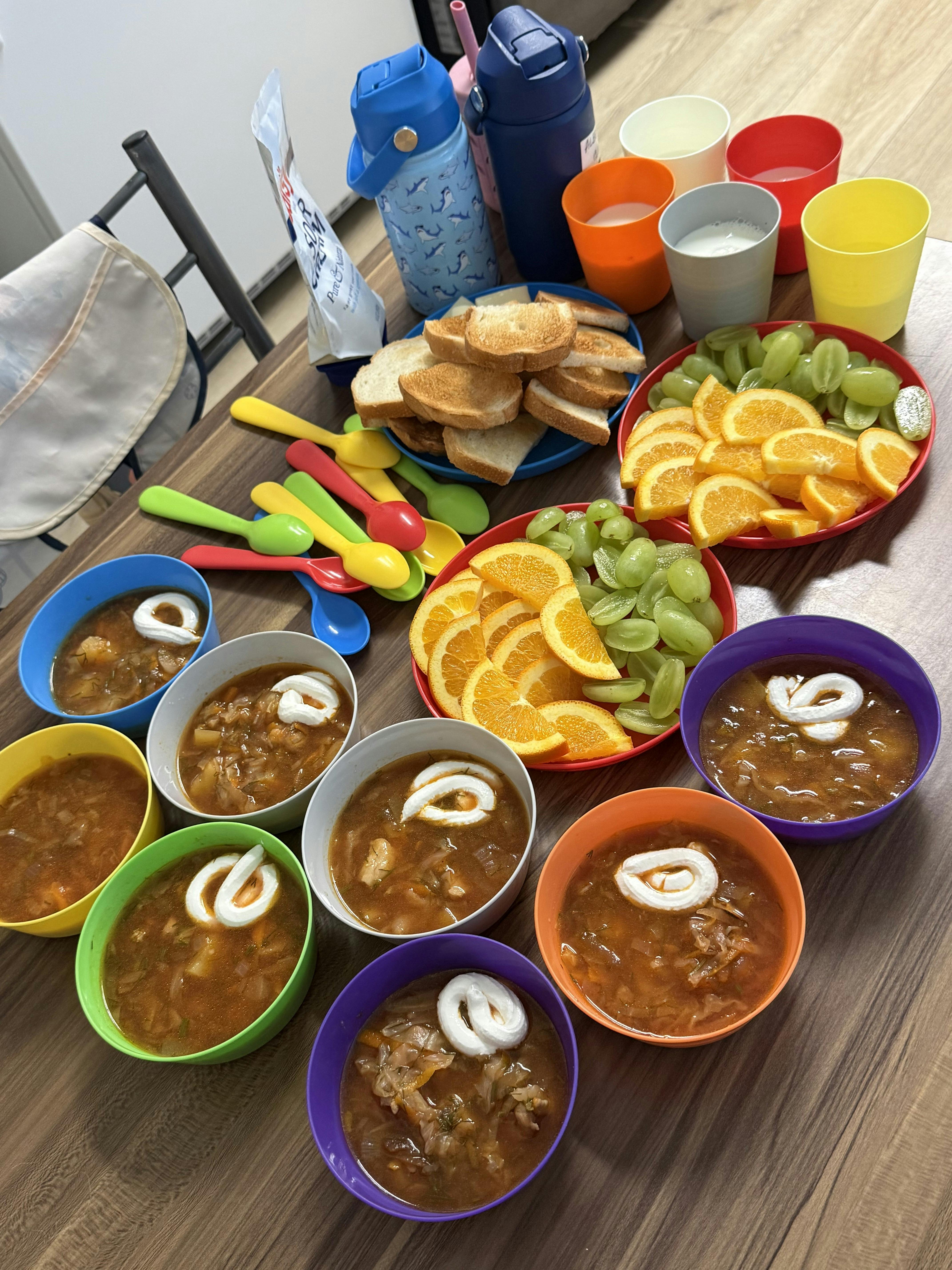 Multiple bowls of soup with sour cream, sliced fruit, and toast on a table