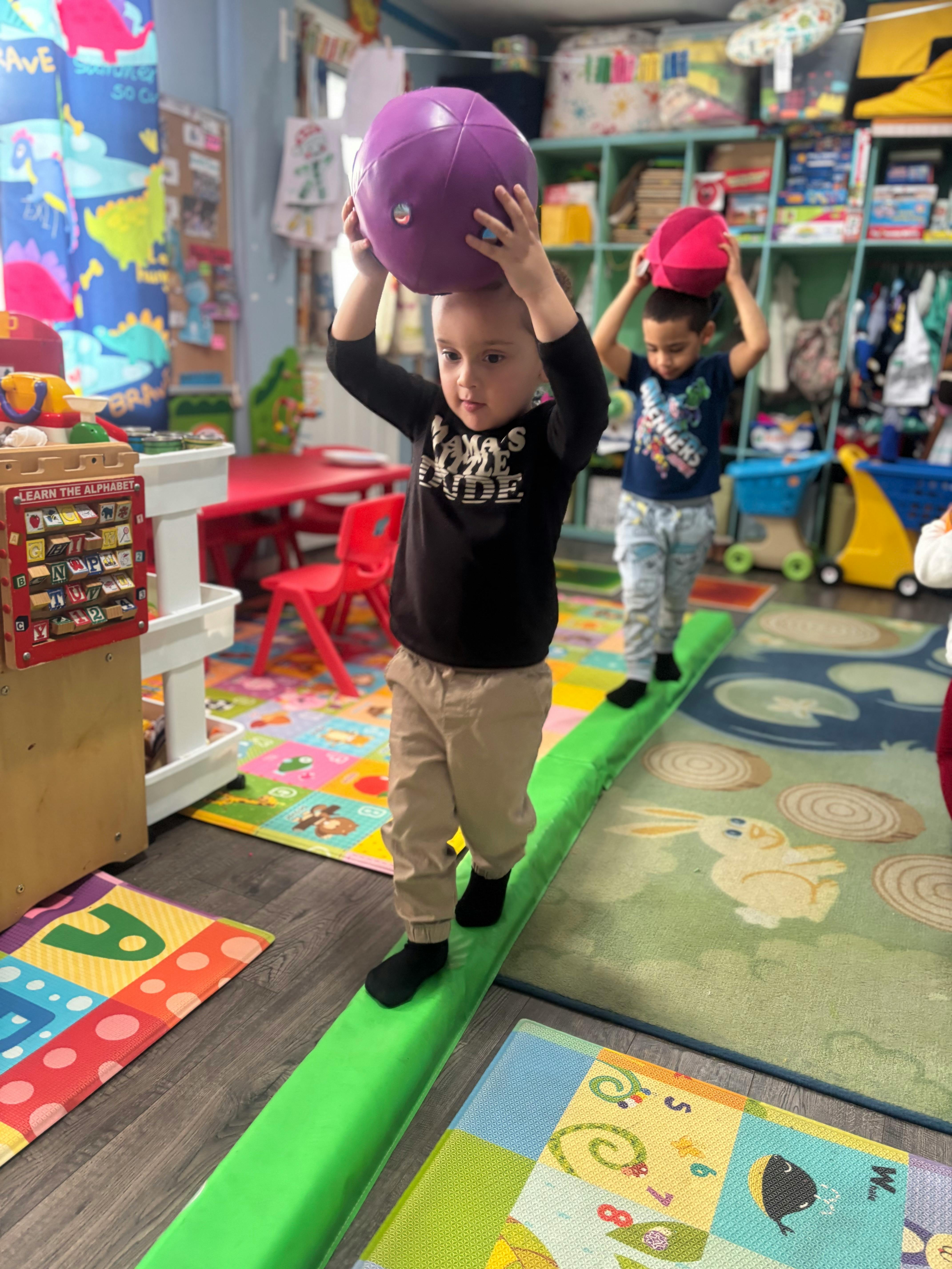 Two children balancing on a green beam, holding balls above their heads