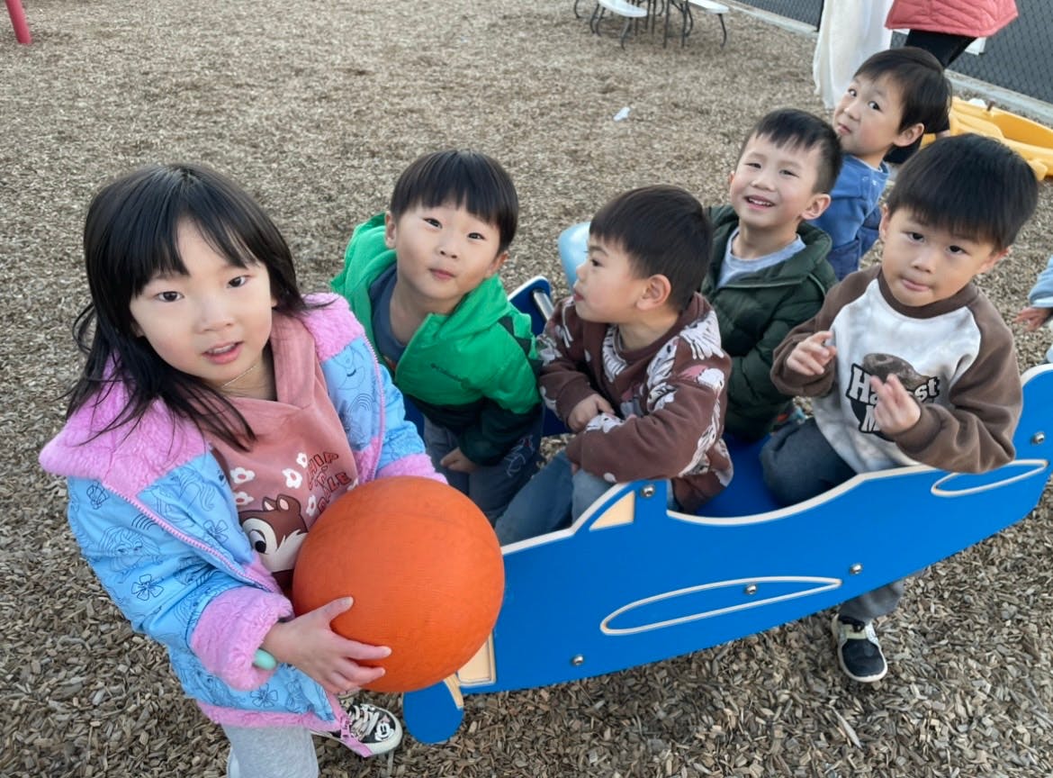 Children playing in a blue airplane-shaped toy, one child holding an orange ball
