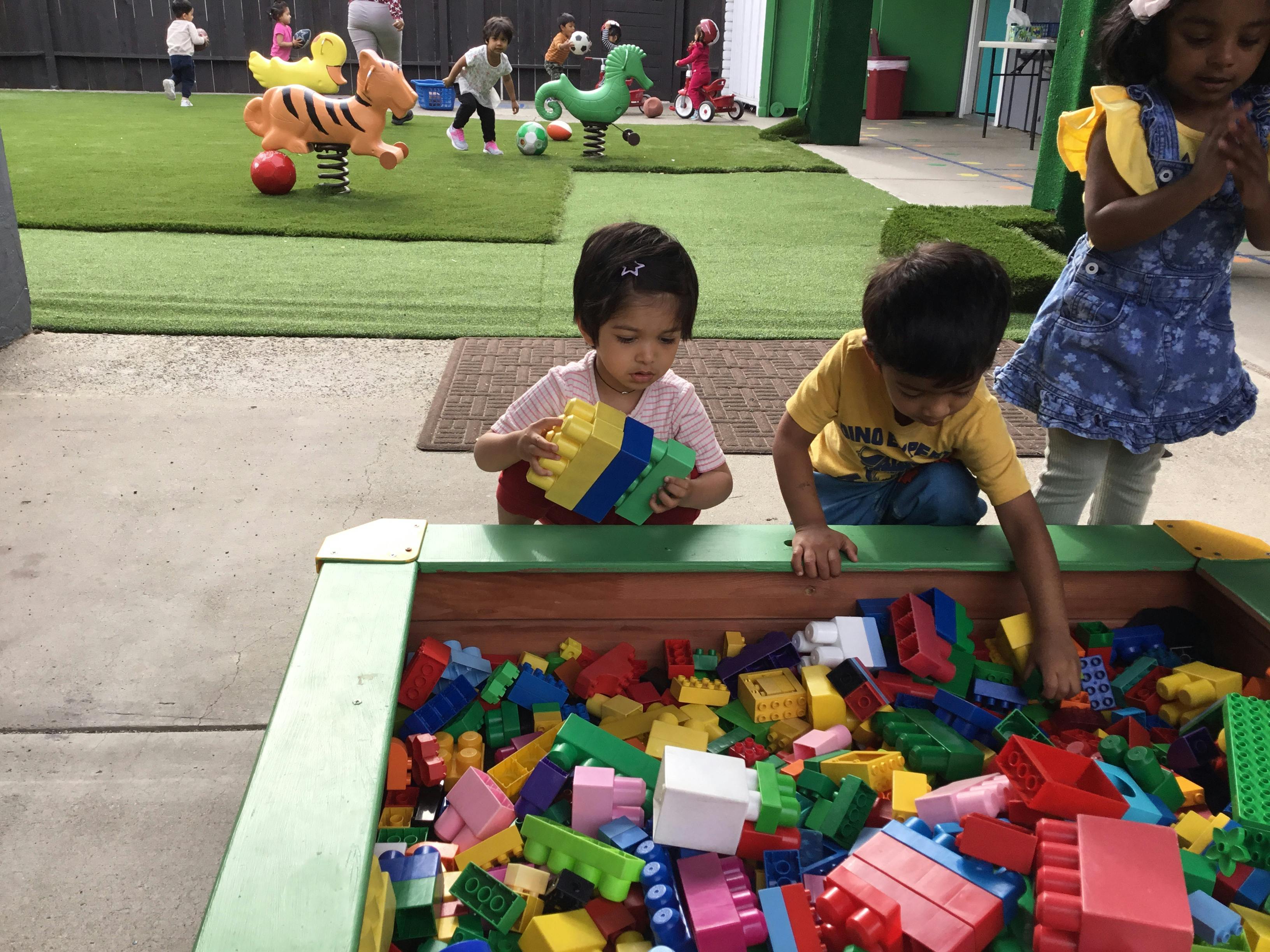 Children playing with large building blocks in a sandbox with a playground in background