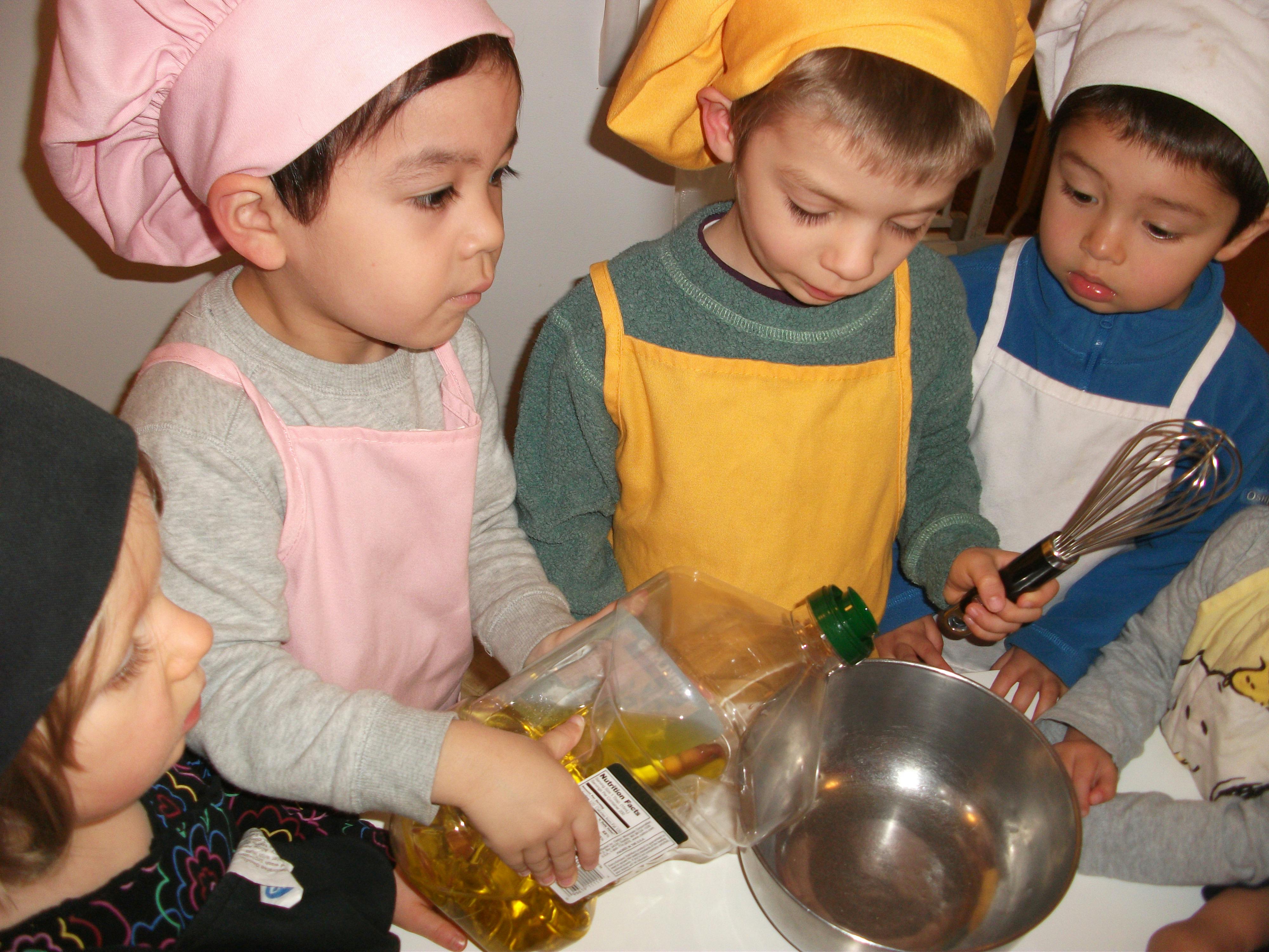 Children in chef hats and aprons, one pouring liquid from a bottle into a bowl