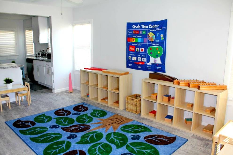 Classroom with a blue rug, shelves with learning materials, and a wall chart