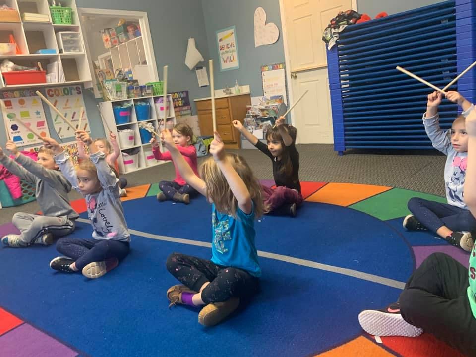 Children sitting on a mat with arms raised holding sticks