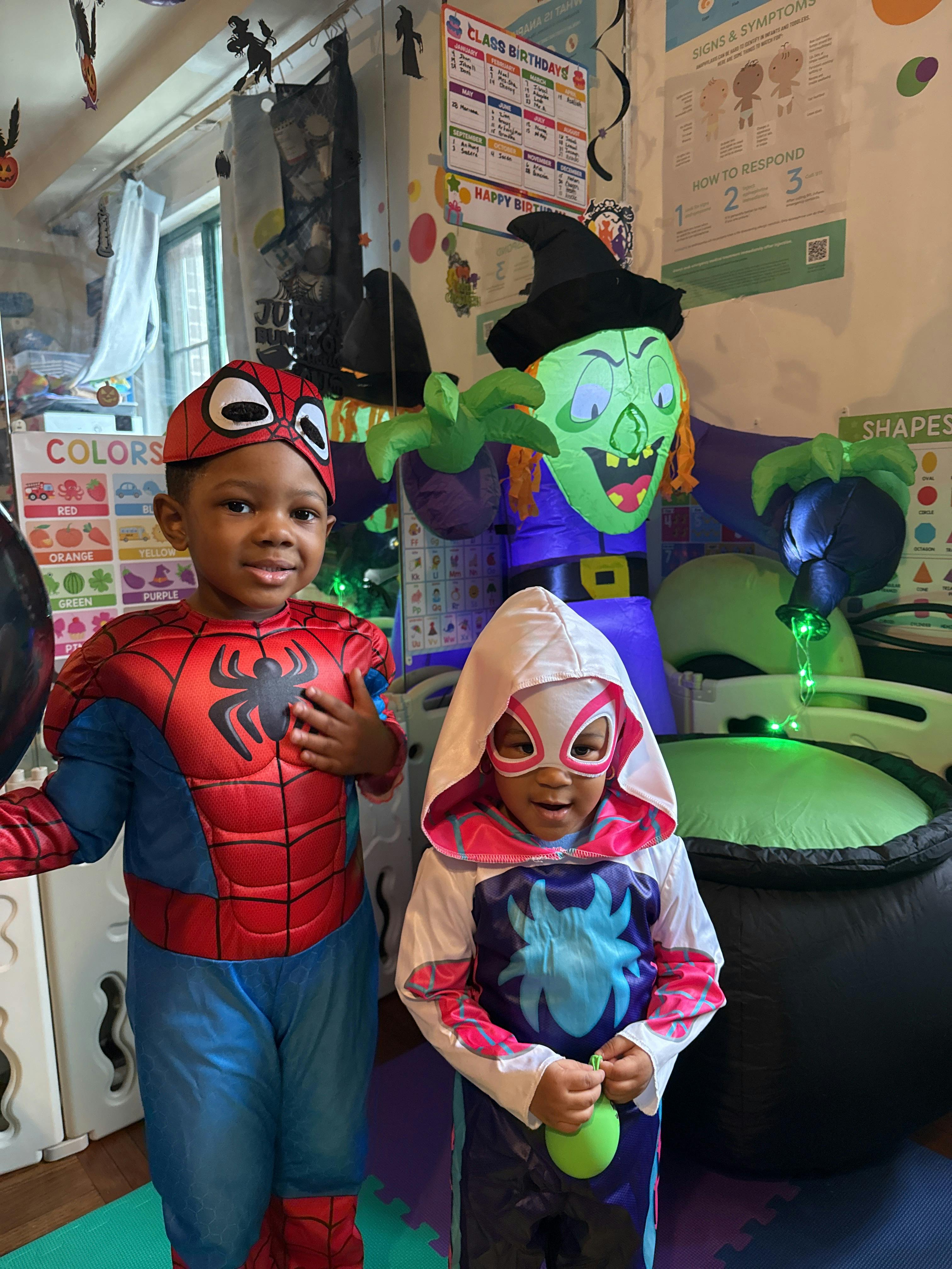 Two children in superhero costumes standing in front of Halloween decorations