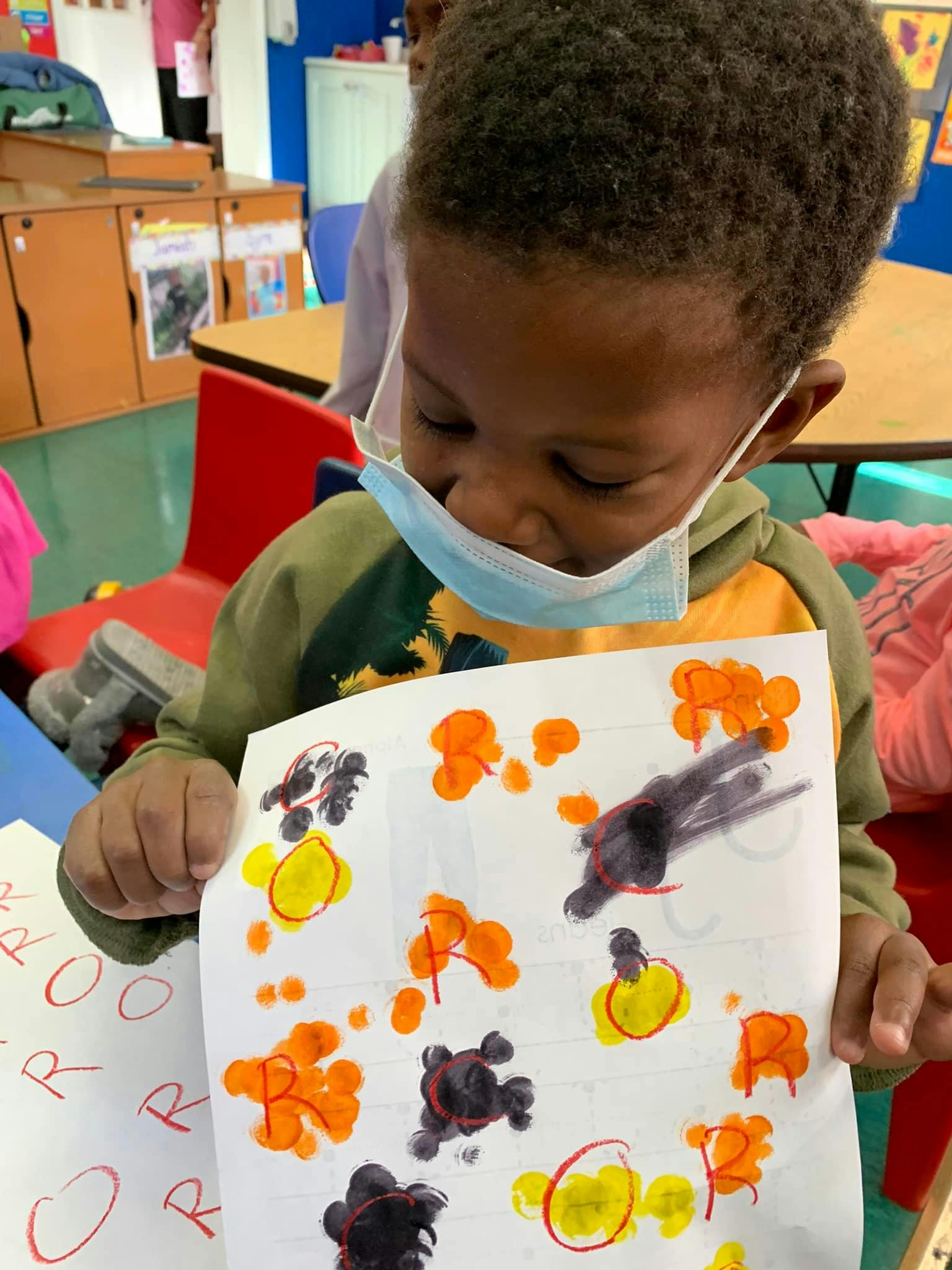 Child in a mask holding up a paper with colorful paint splotches