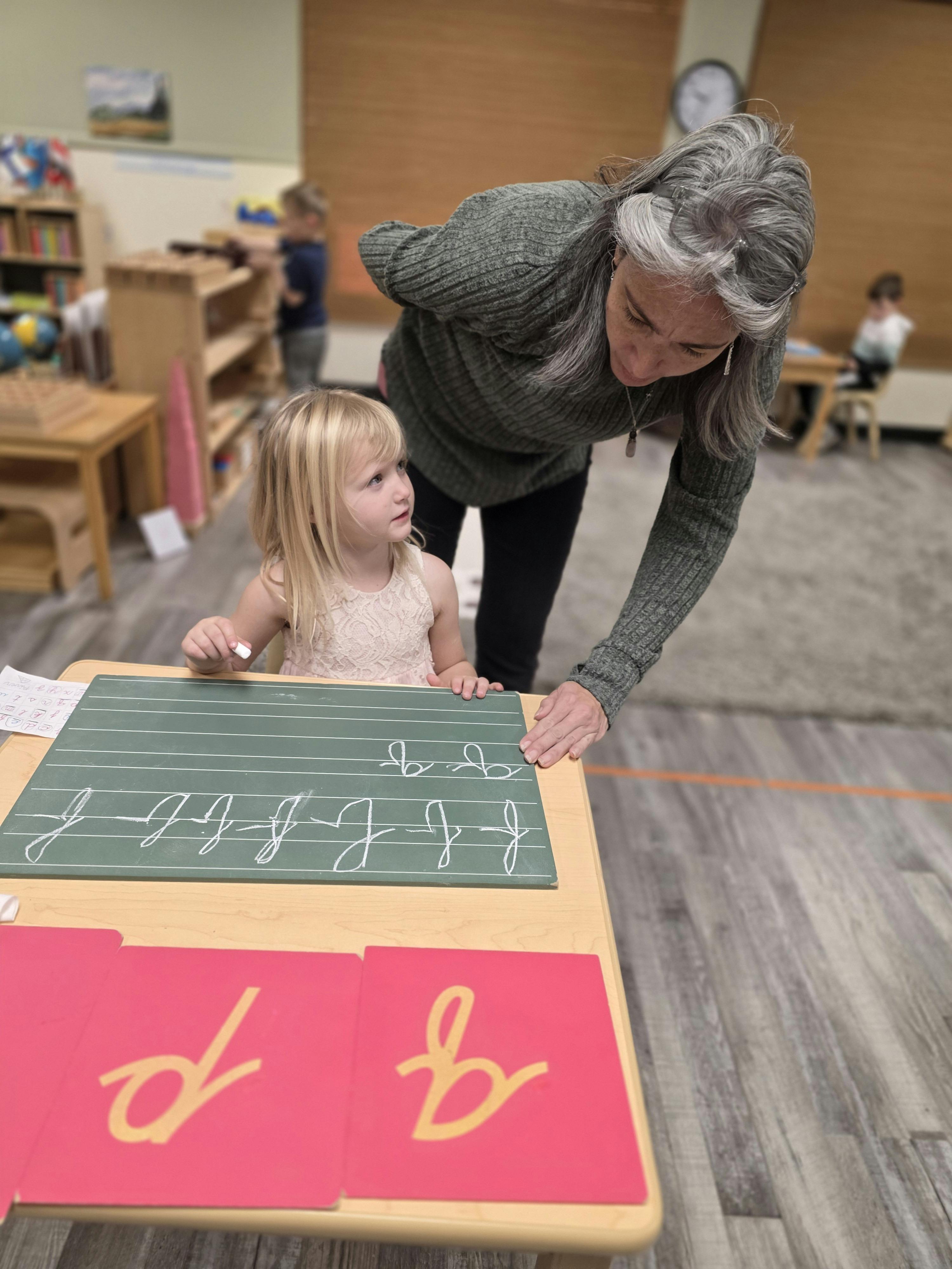 Adult helping a child write letters on a chalkboard in a classroom