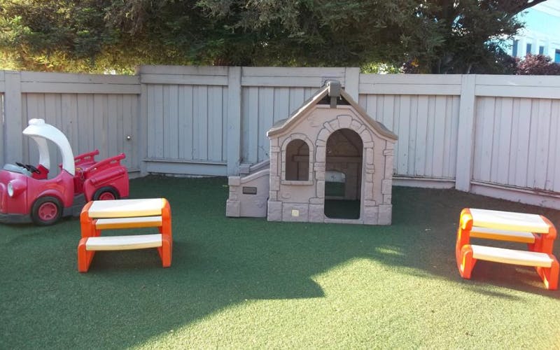 A beige playhouse, two red toy cars, and two orange benches on artificial grass