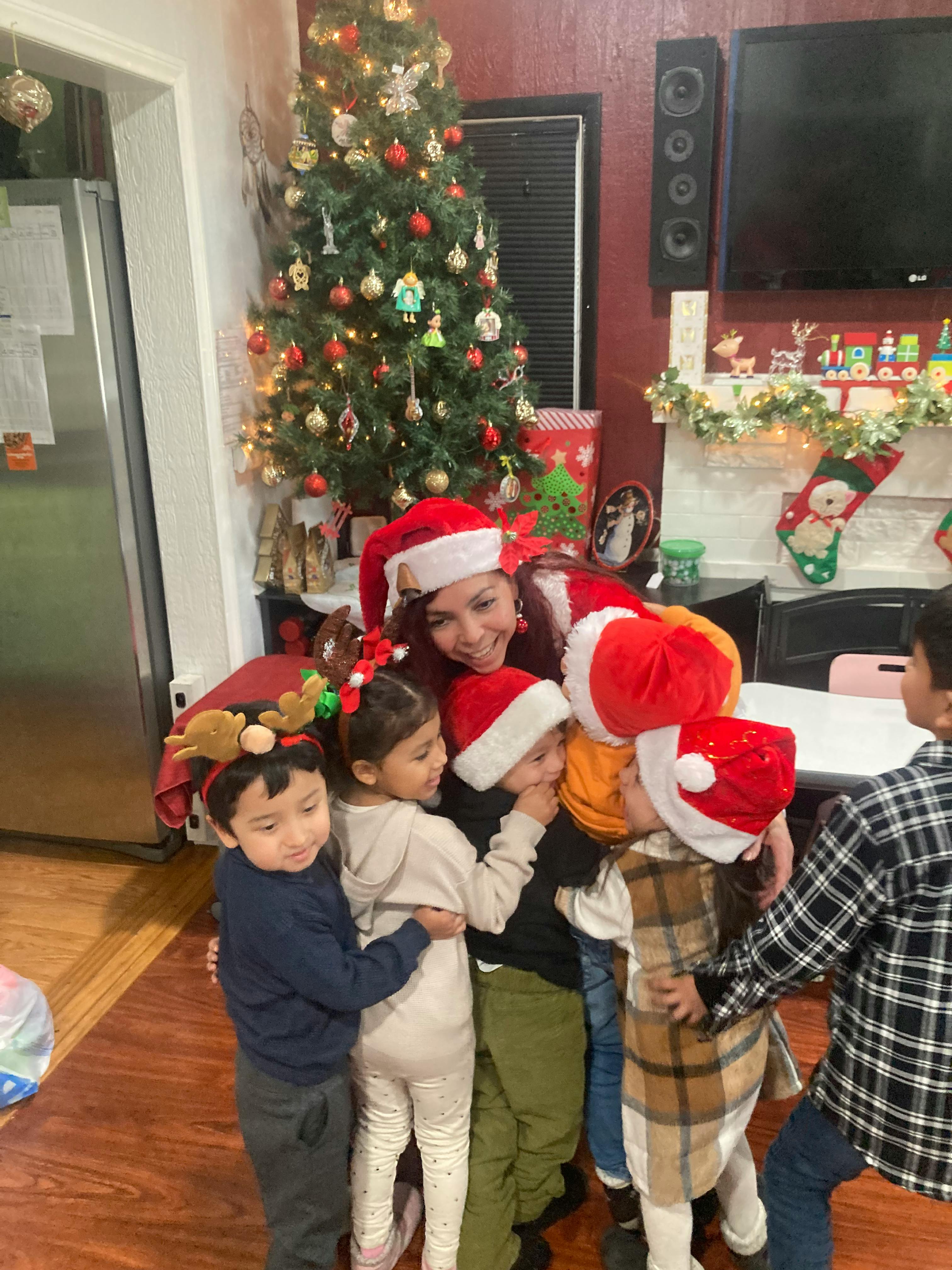 A person wearing a Santa hat hugging a group of children by a Christmas tree