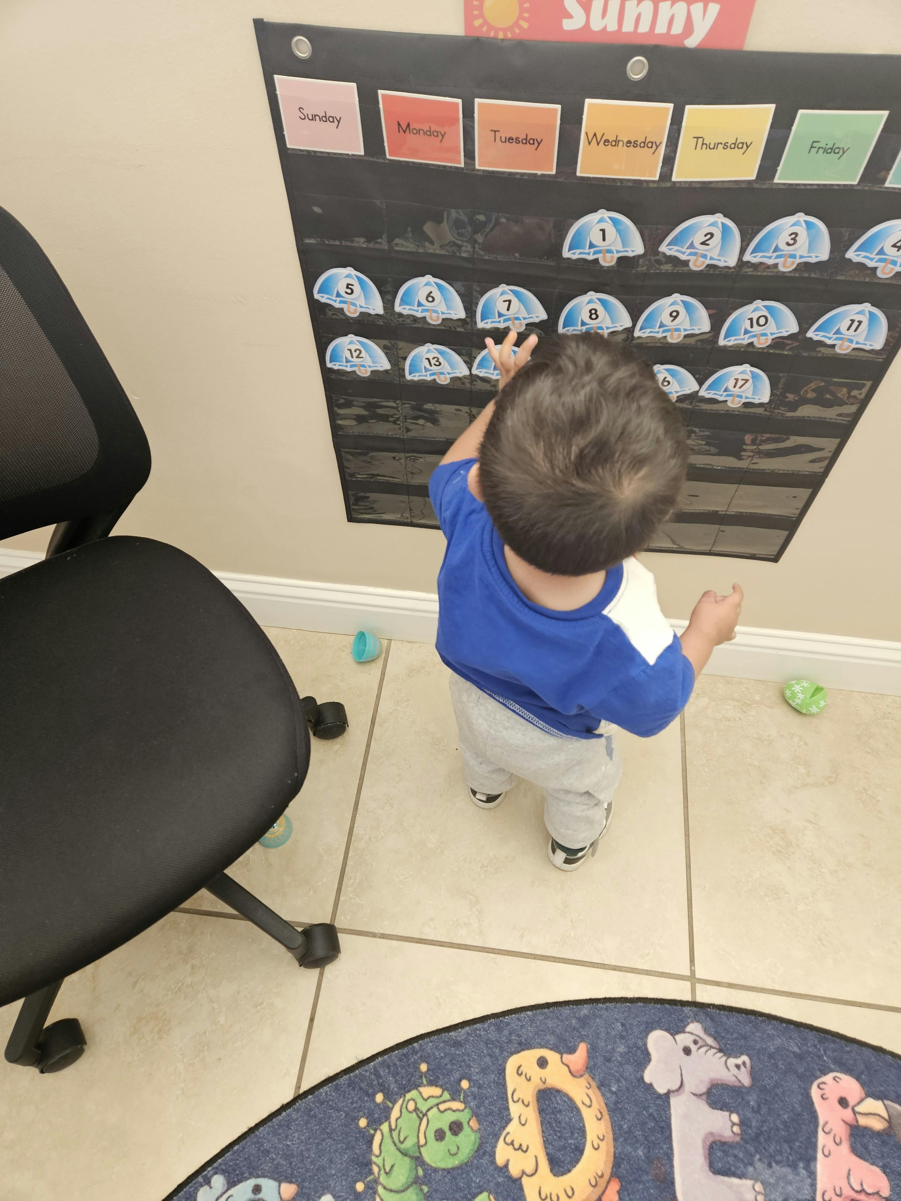 Child reaching towards a wall calendar with days of the week and numbered umbrellas
