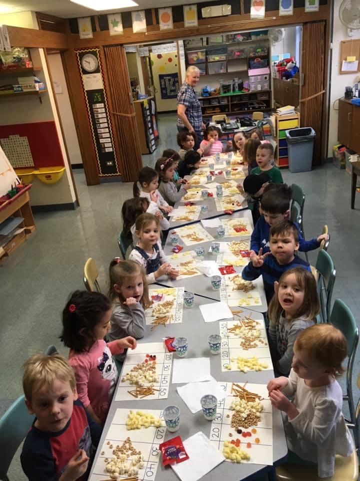 Children sitting at tables in a classroom with an adult standing in the background