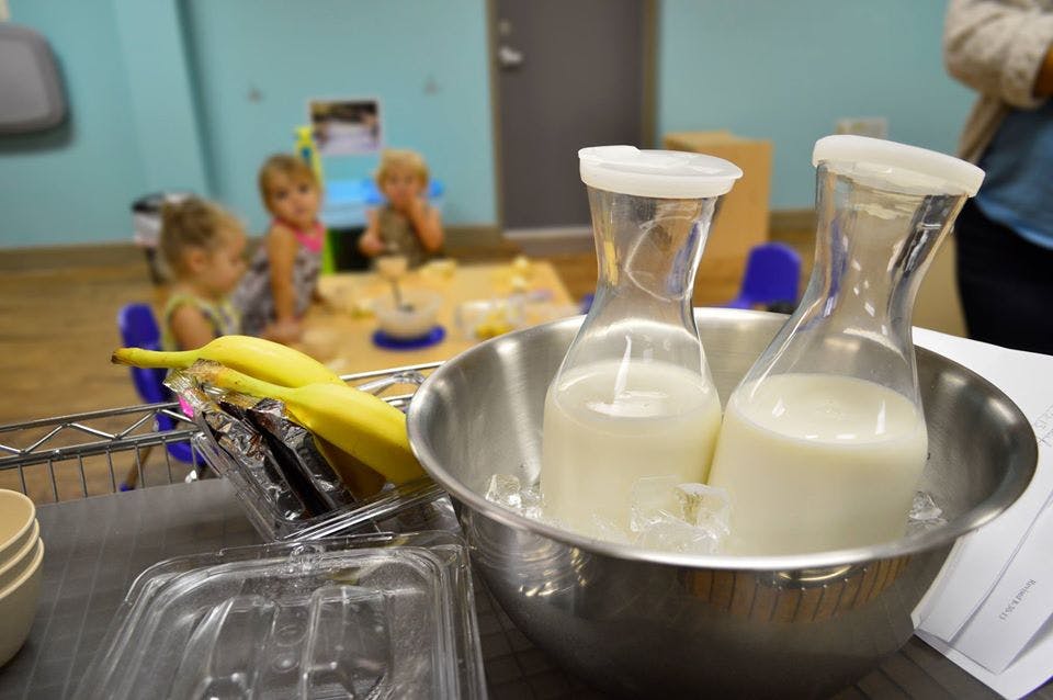Two carafes of milk in a bowl with bananas and children in the background