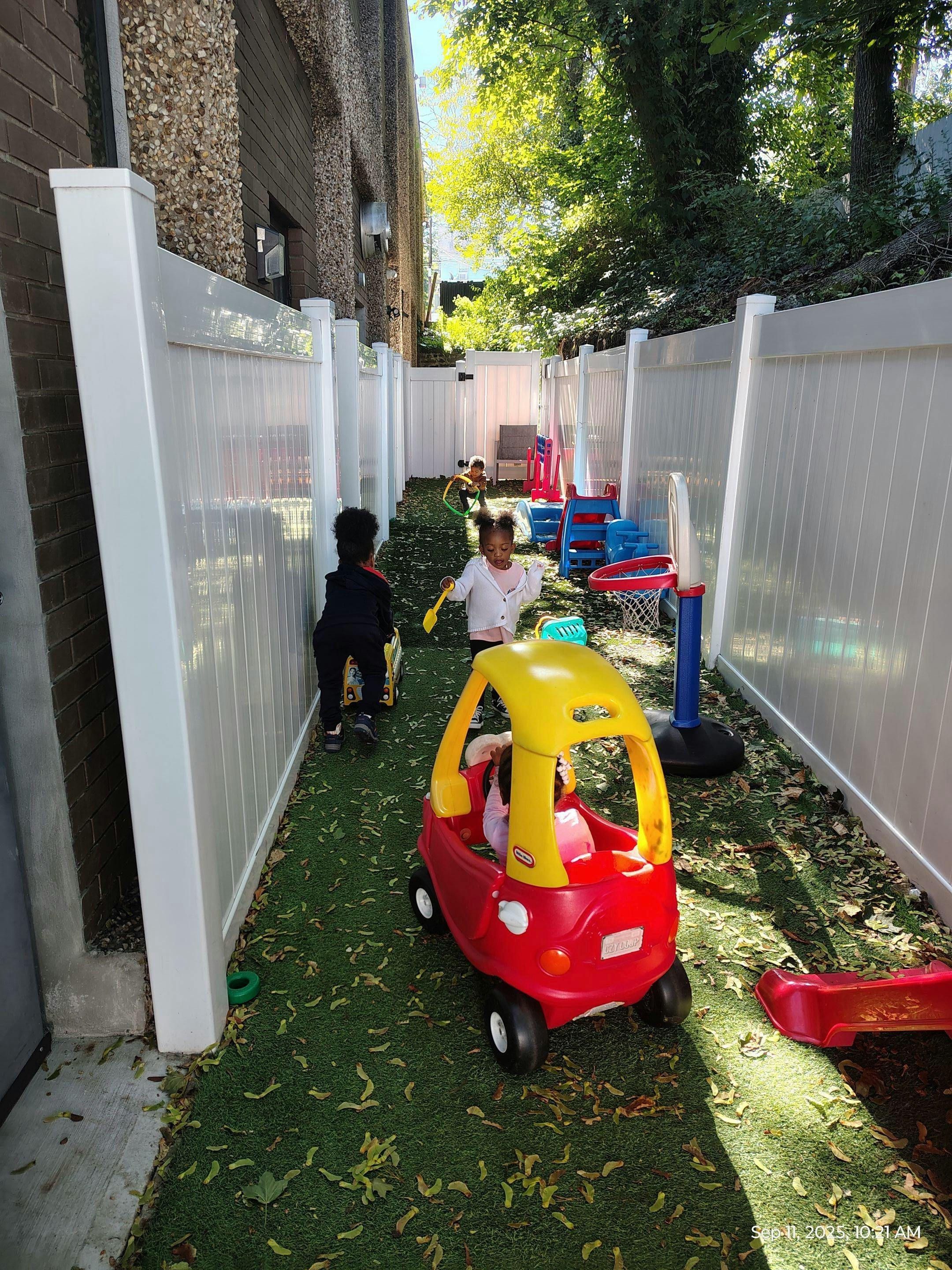 Children playing in a fenced outdoor area with toy cars and a basketball hoop