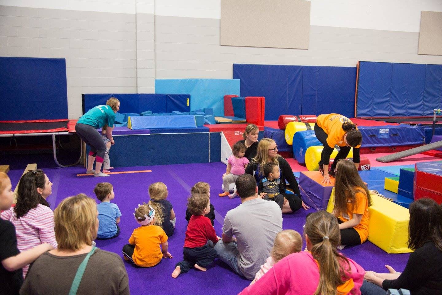 Adults and children on a purple mat in a gym with blue and yellow equipment