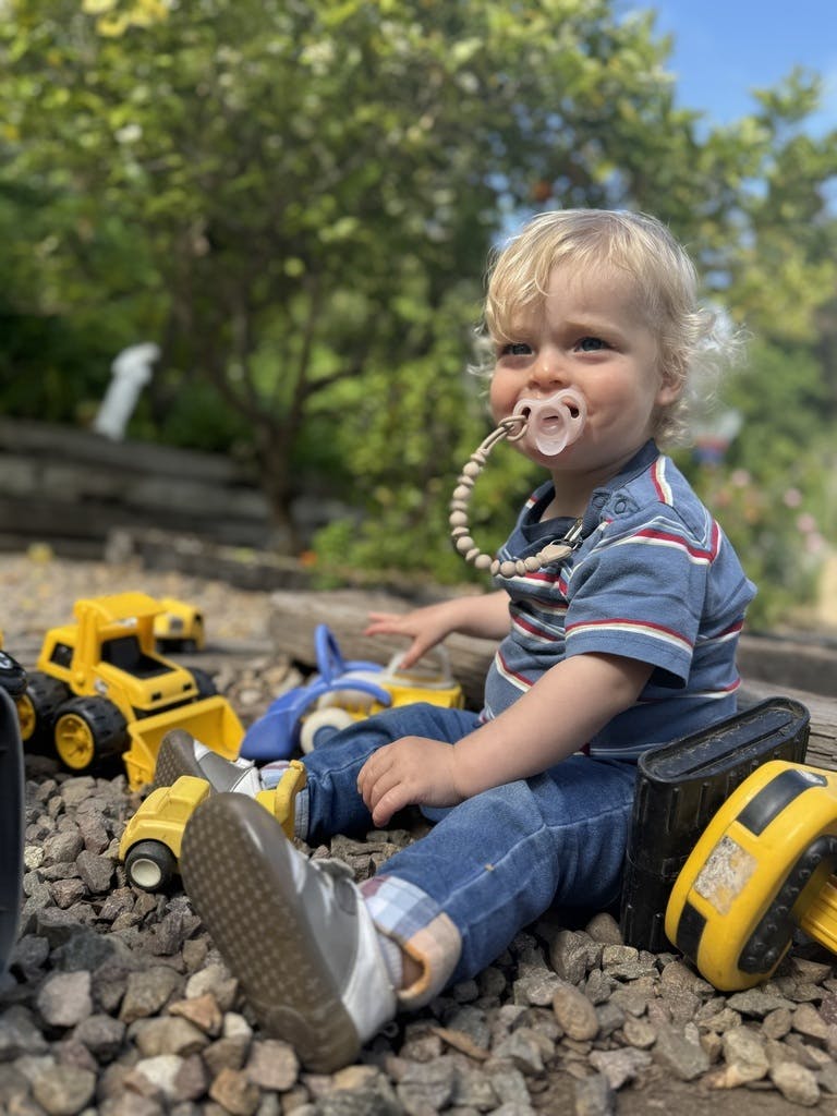 Child with pacifier sitting on rocks with toy trucks