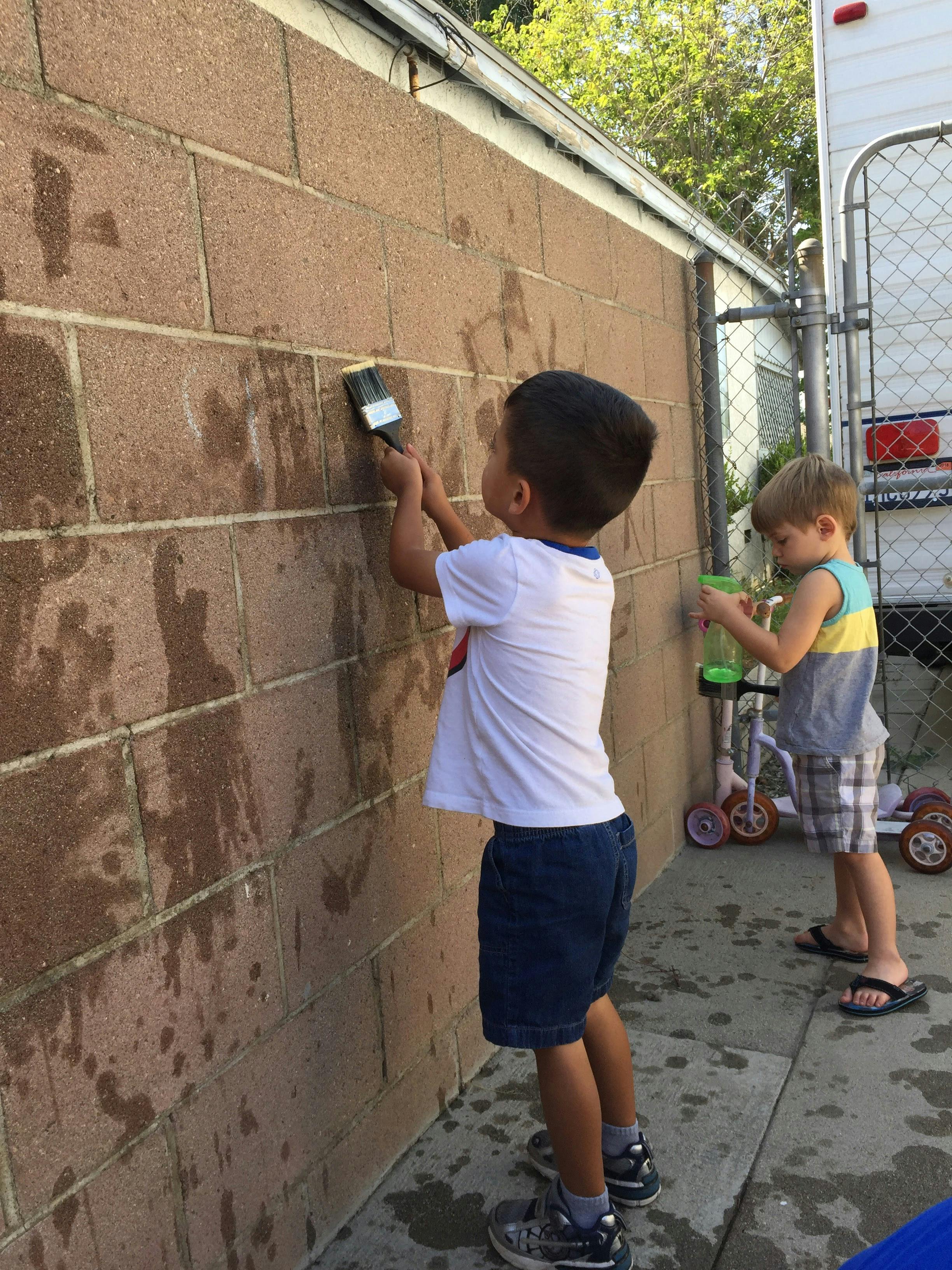 Child painting a brick wall with a brush, another child nearby