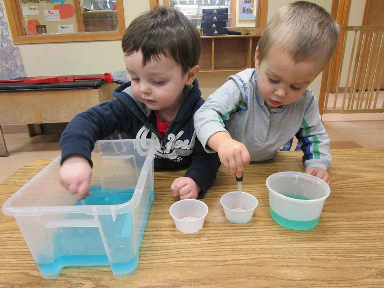 Two children playing with water and cups on a wooden surface