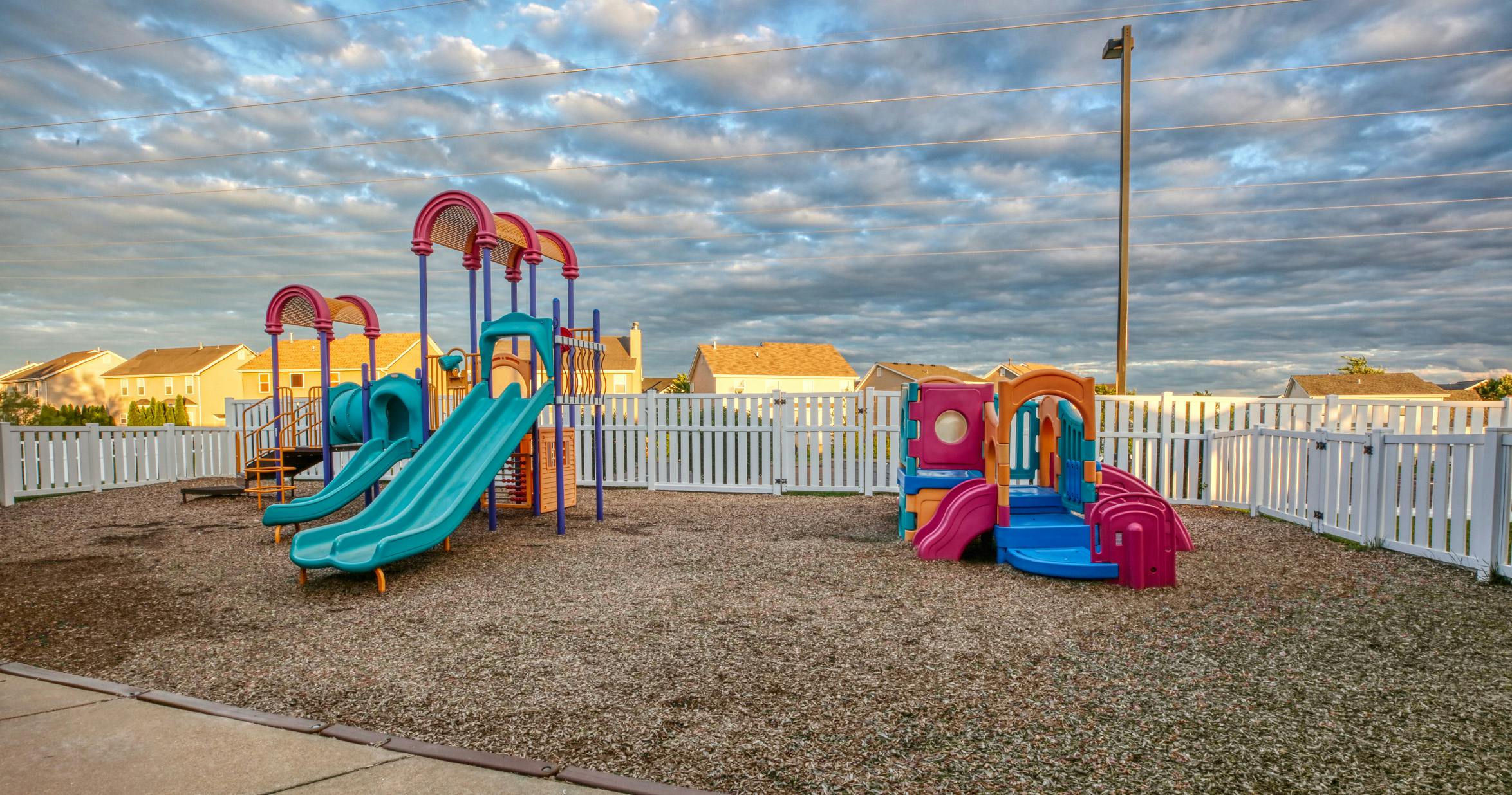 A colorful playground with slides and a tunnel on wood chips, fenced by white picket