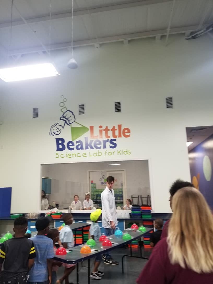 Children and adults in a science lab with "Little Beakers Science Lab for Kids" sign