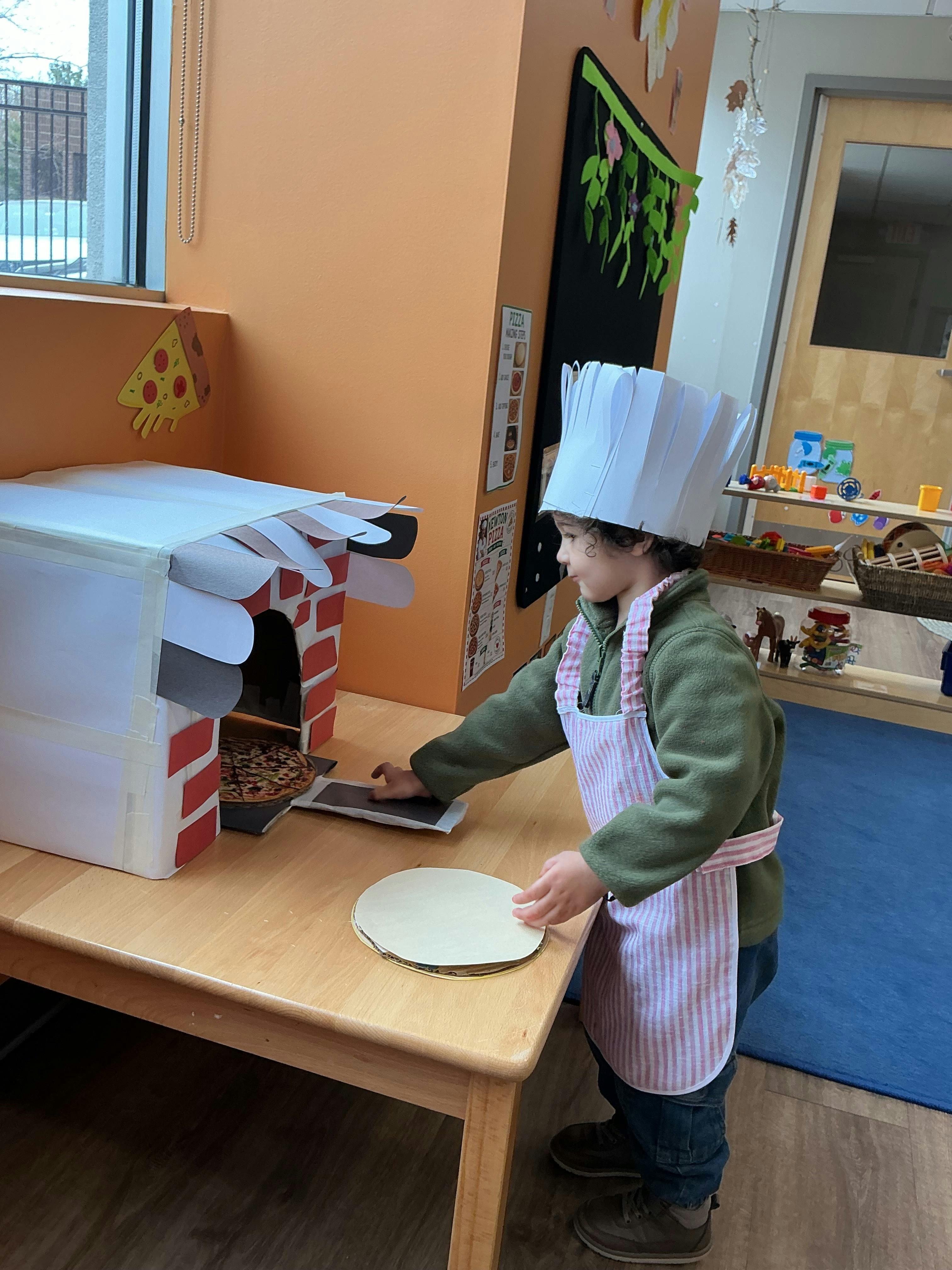 Child wearing chef hat and apron, putting toy pizza in a play oven