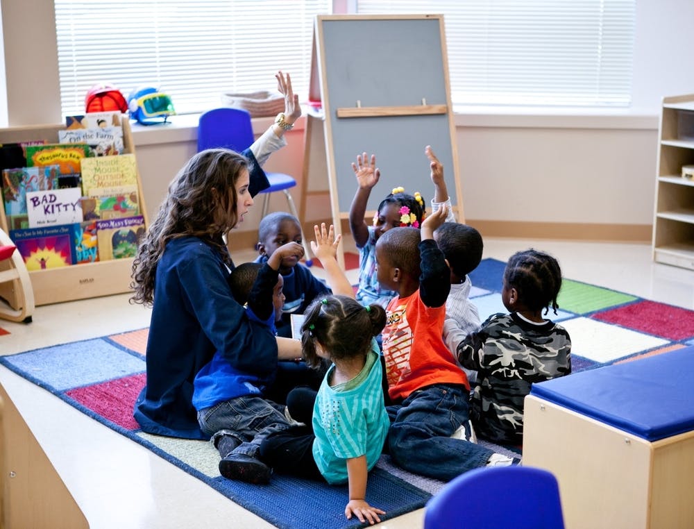 Adult and children sitting on a rug with hands raised in a classroom