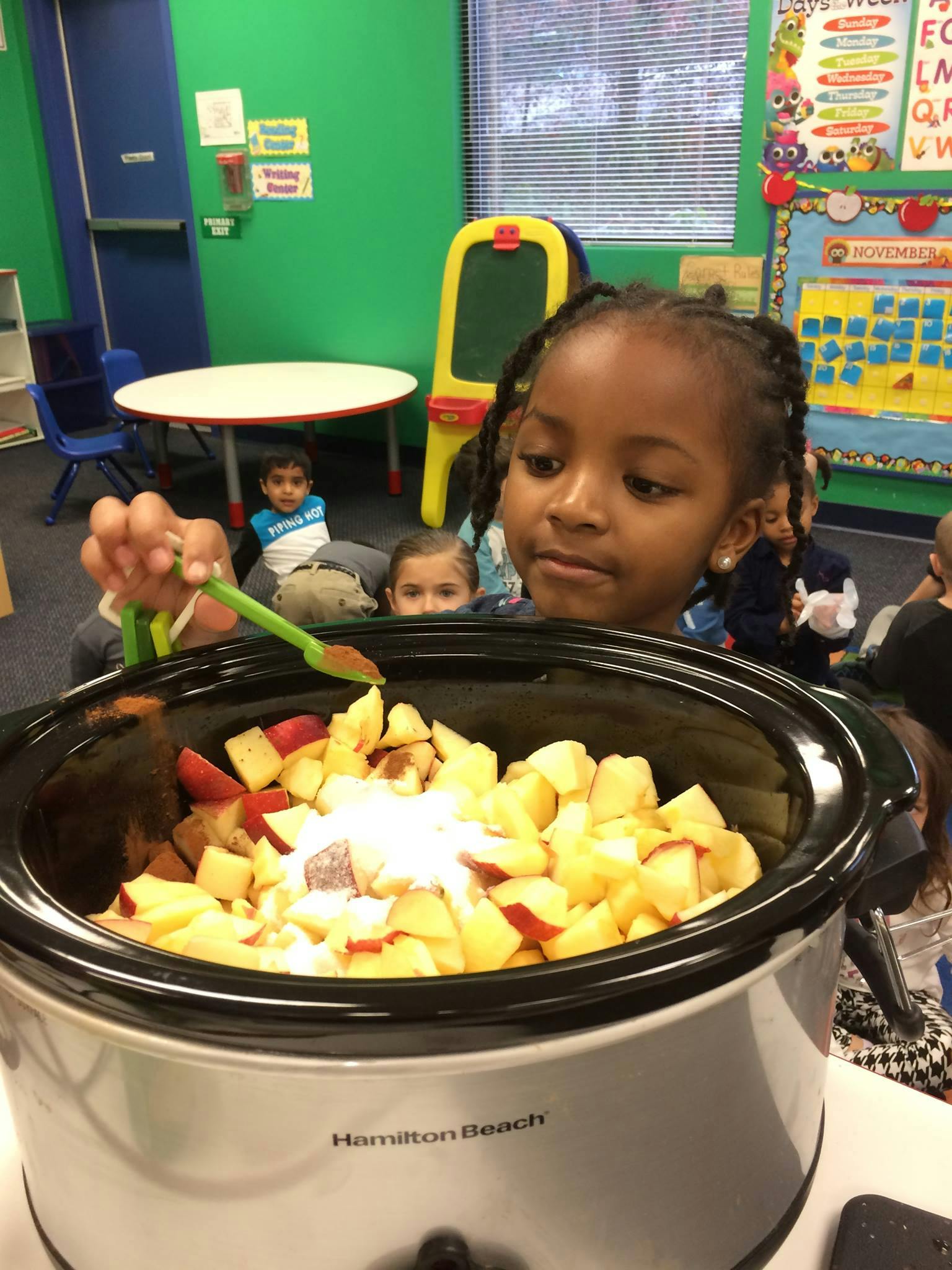 Child adding ingredients to a slow cooker filled with chopped apples in a classroom