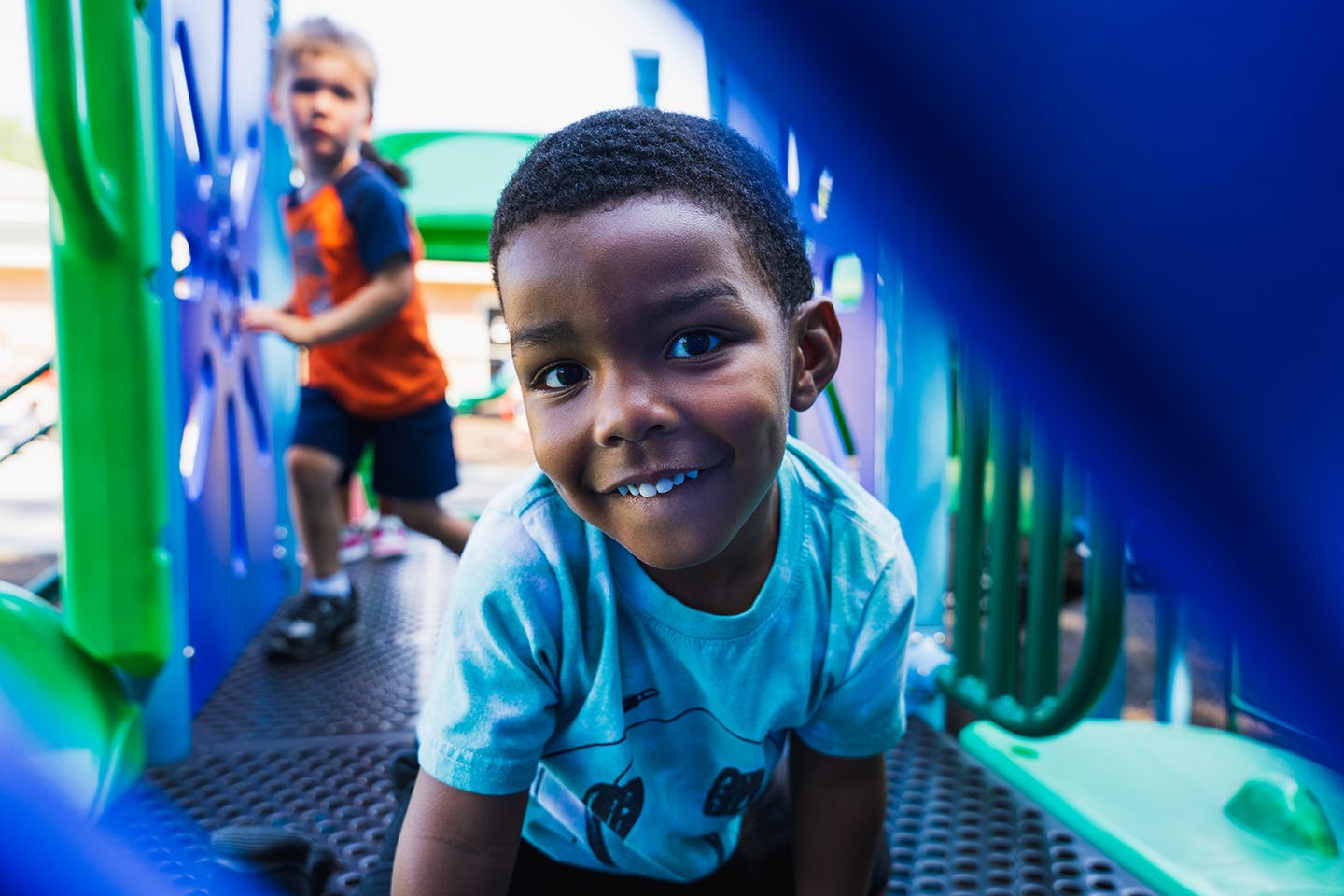 Child on playground equipment looking at camera, another child in background