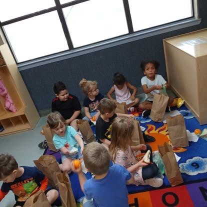 Group of children sitting on a colorful carpet in a classroom with lunch bags
