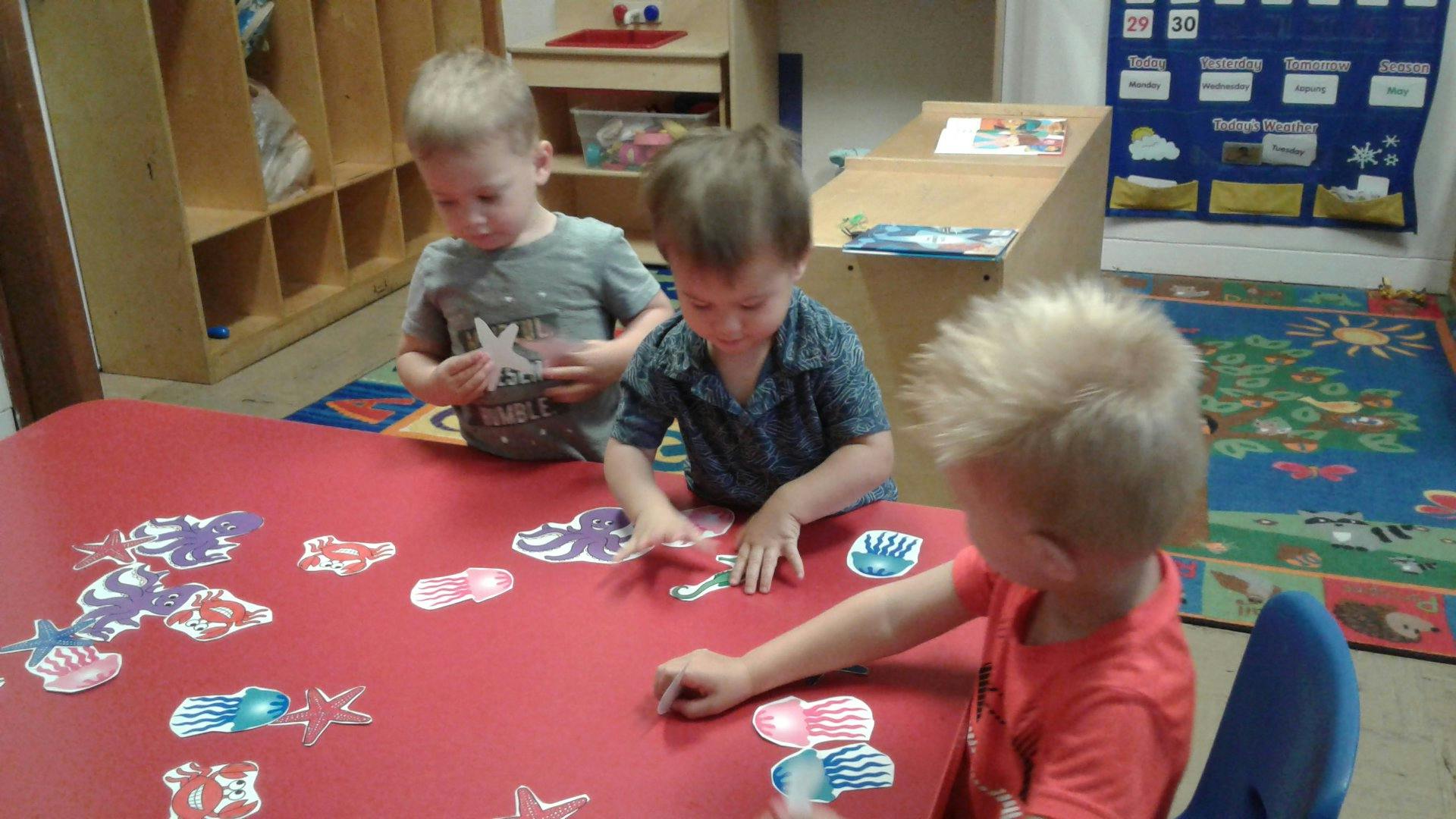 Three children playing with cutouts at a red table