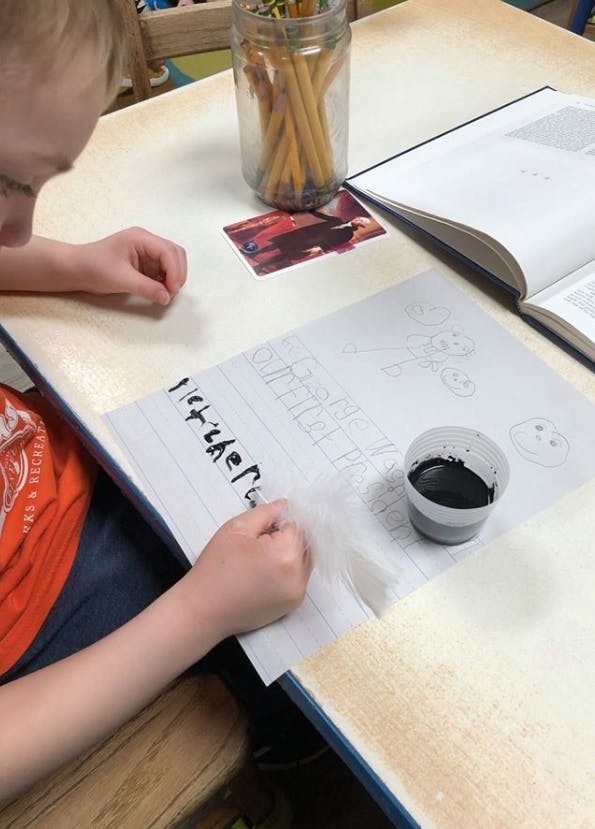 Child writing on paper with a cup of ink on a table