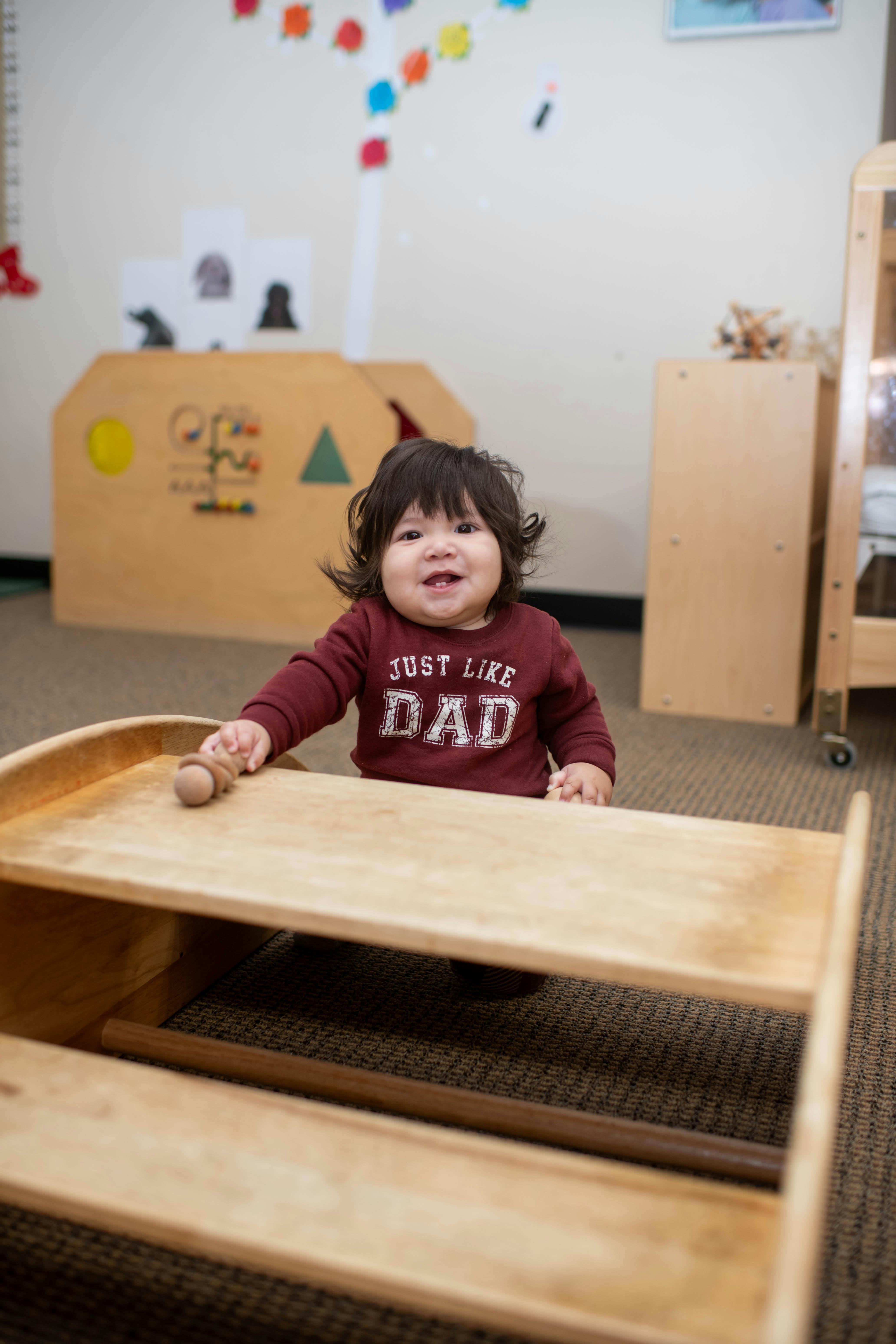 Child in a burgundy sweatshirt that says "JUST LIKE DAD" standing behind a wooden cart
