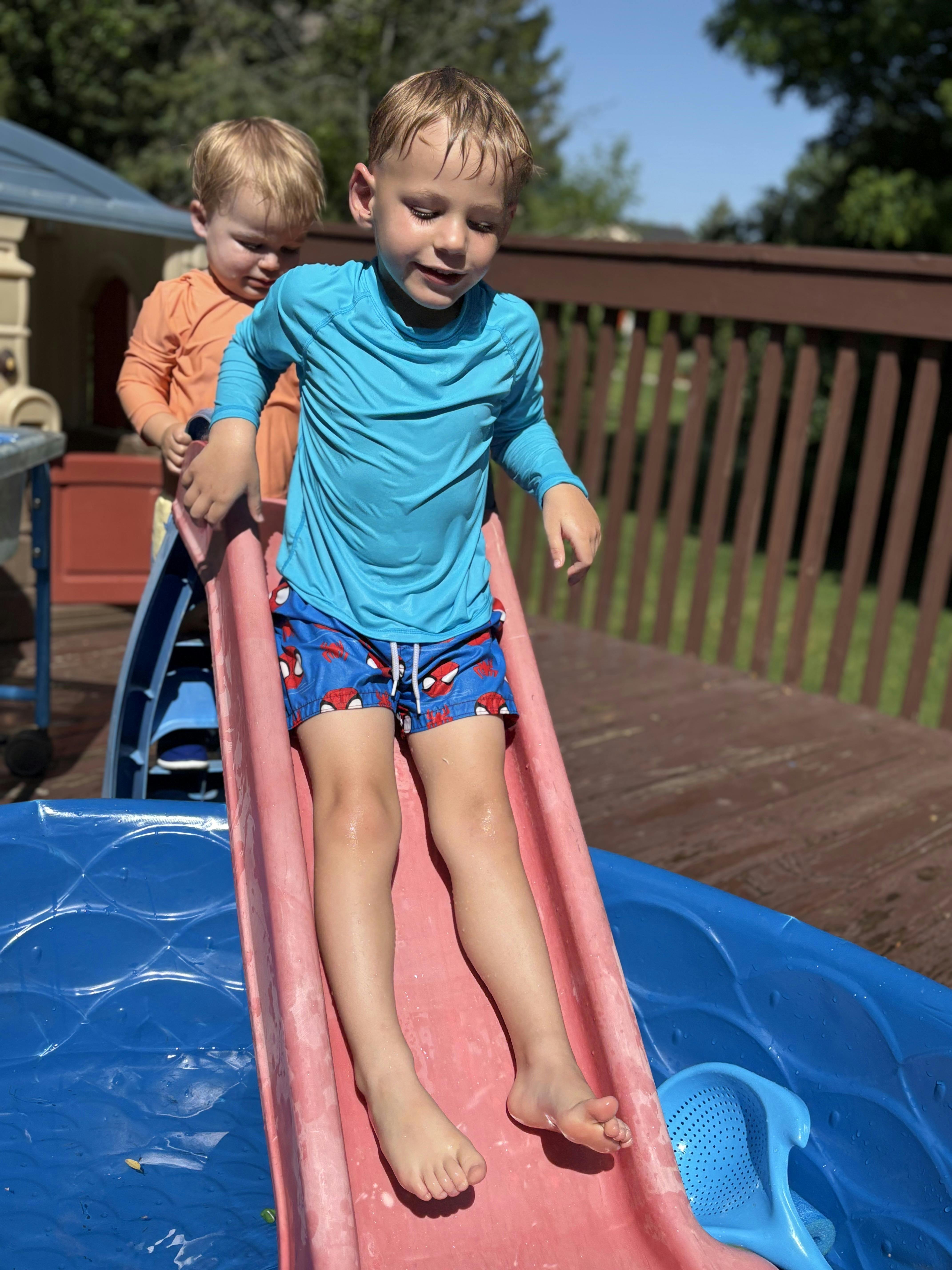 Child in blue shirt and swim trunks going down a slide into a pool