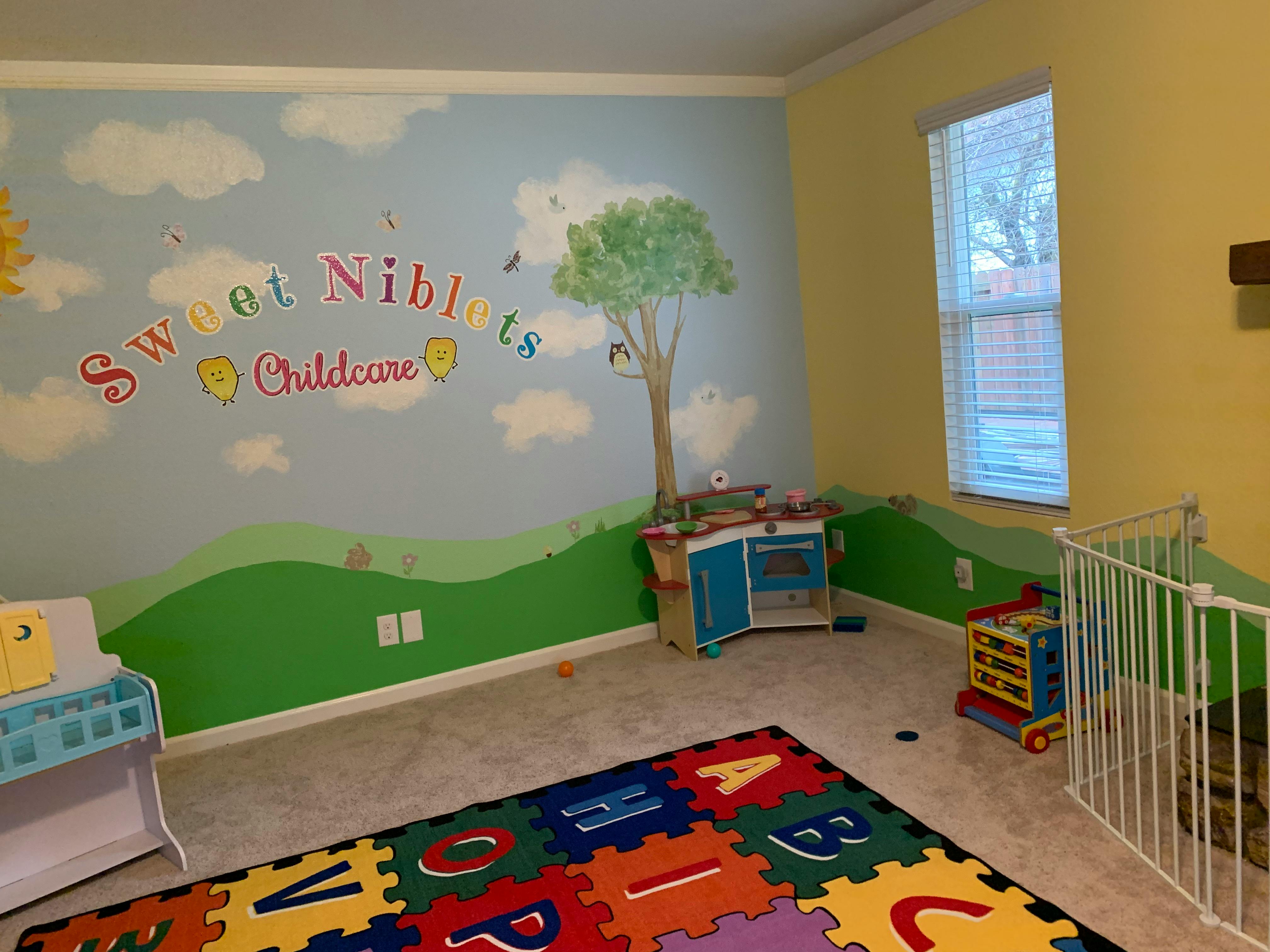 Childcare room with wall mural of sky and grass, play kitchen, and alphabet mat