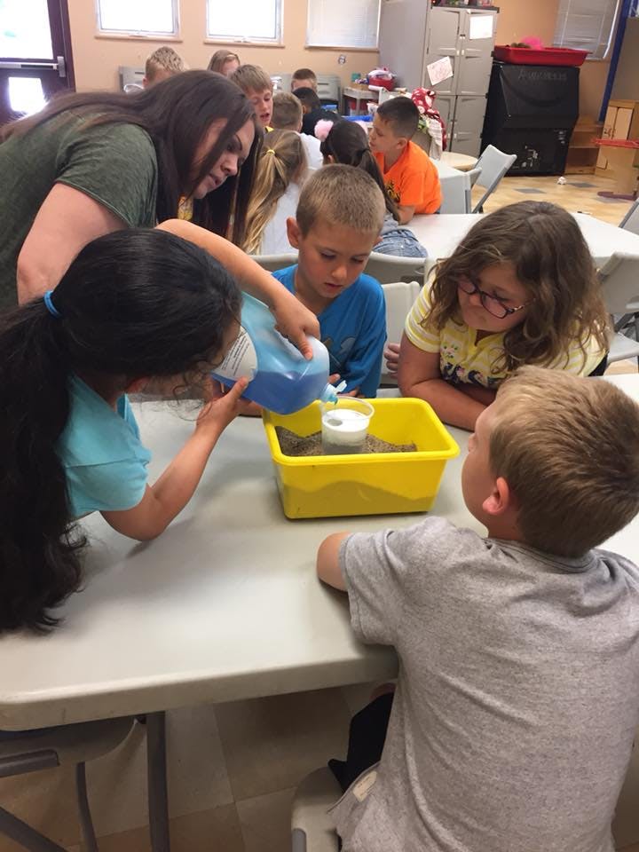 Adult and children gathered around a table, child pouring liquid into a container