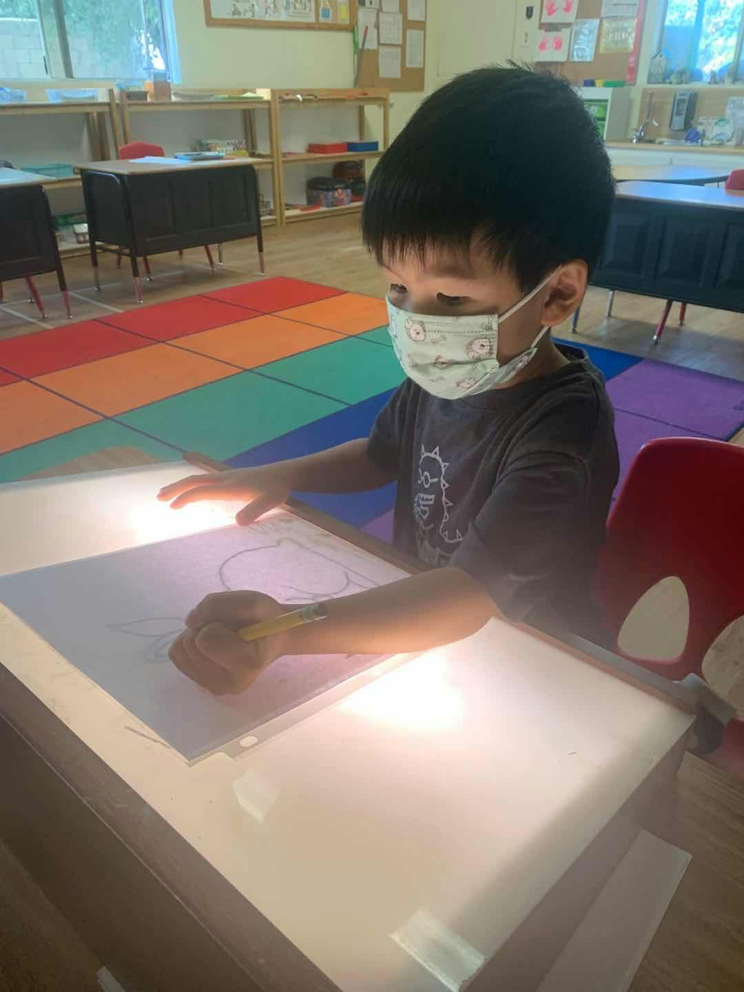 Child in a mask drawing on a light table in a classroom