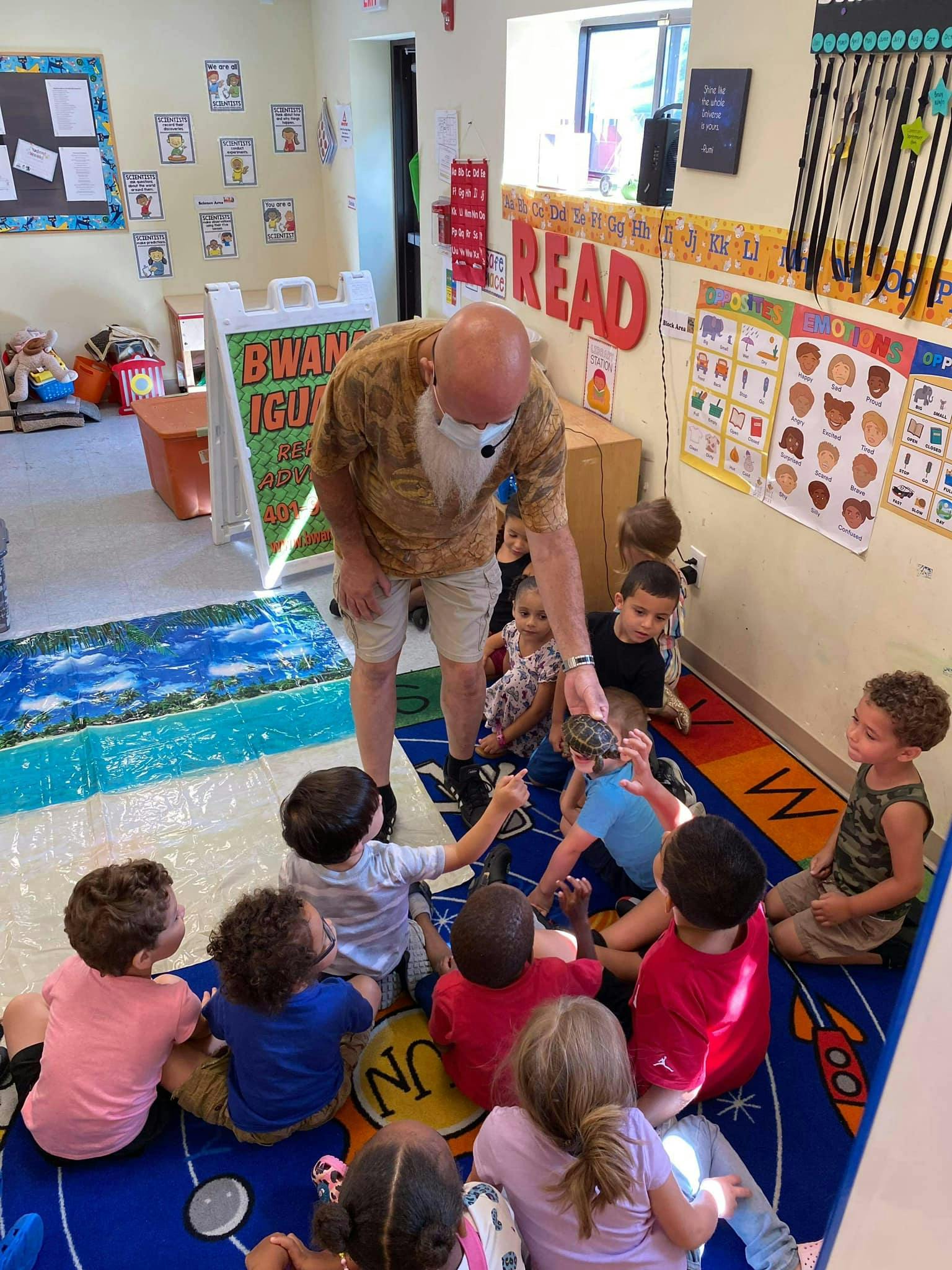 Adult with a white beard showing a small object to children sitting on a rug