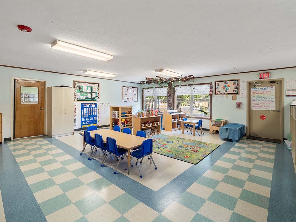Empty classroom with small tables and chairs, and a play rug