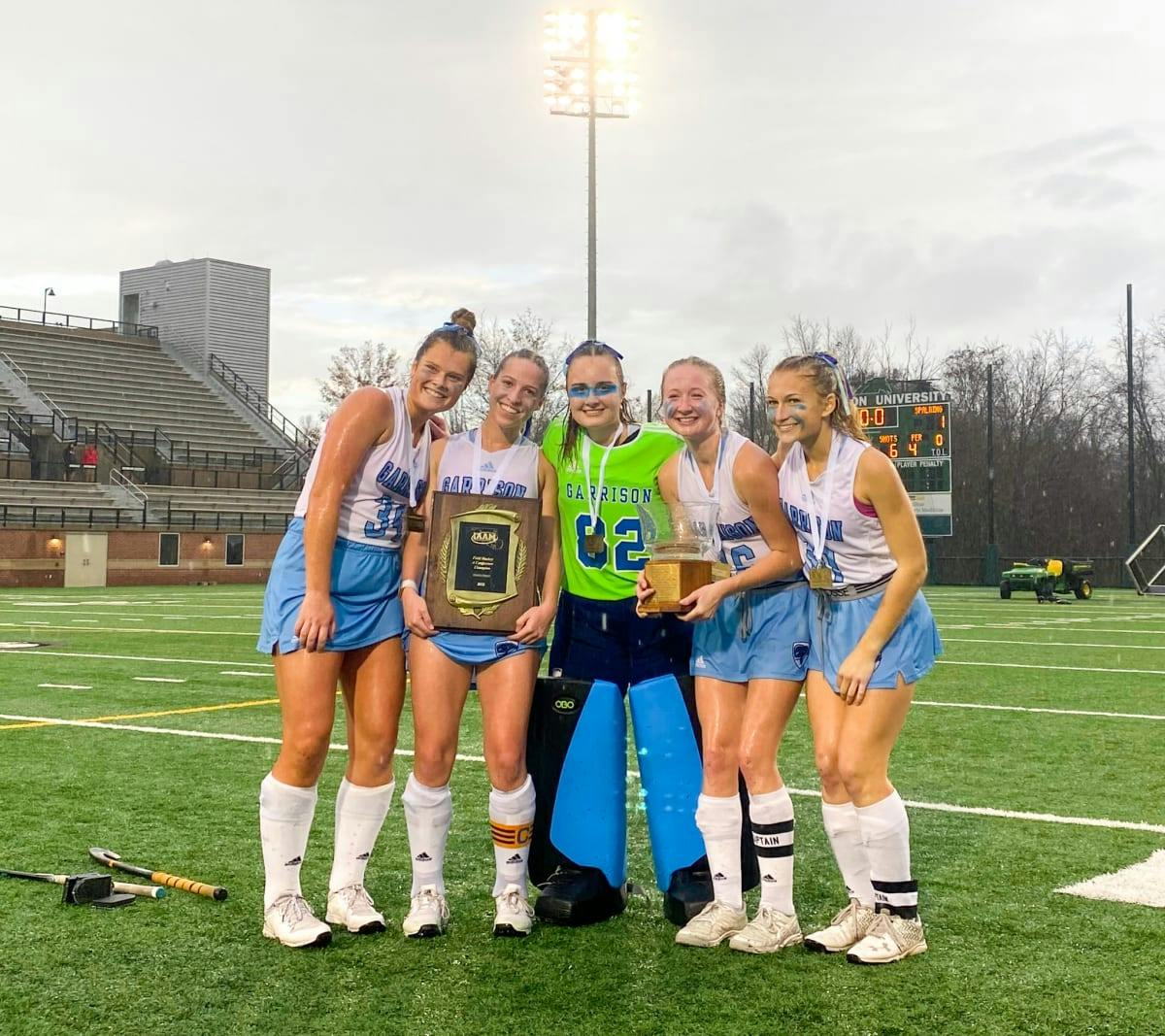 Five people in athletic uniforms holding a trophy and plaque on a sports field