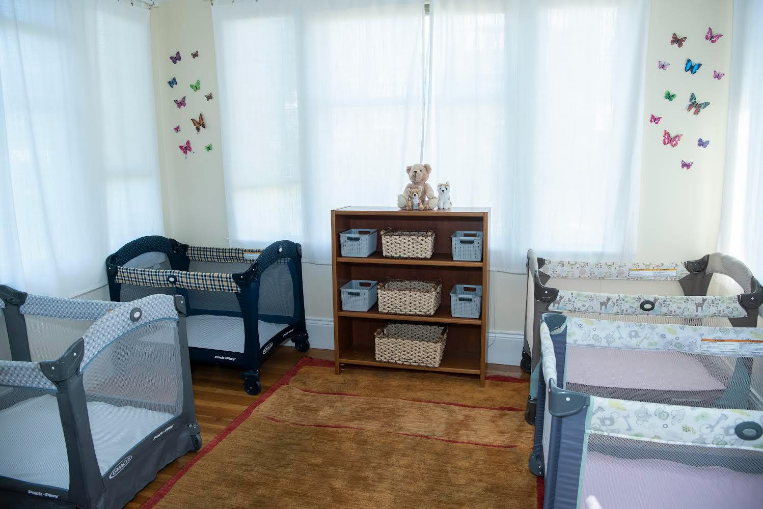Multiple cribs in a room with a bookshelf and butterfly wall decals