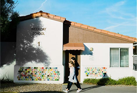 Person and child walking past a building with "WEEKDAY SCHOOL" on the wall