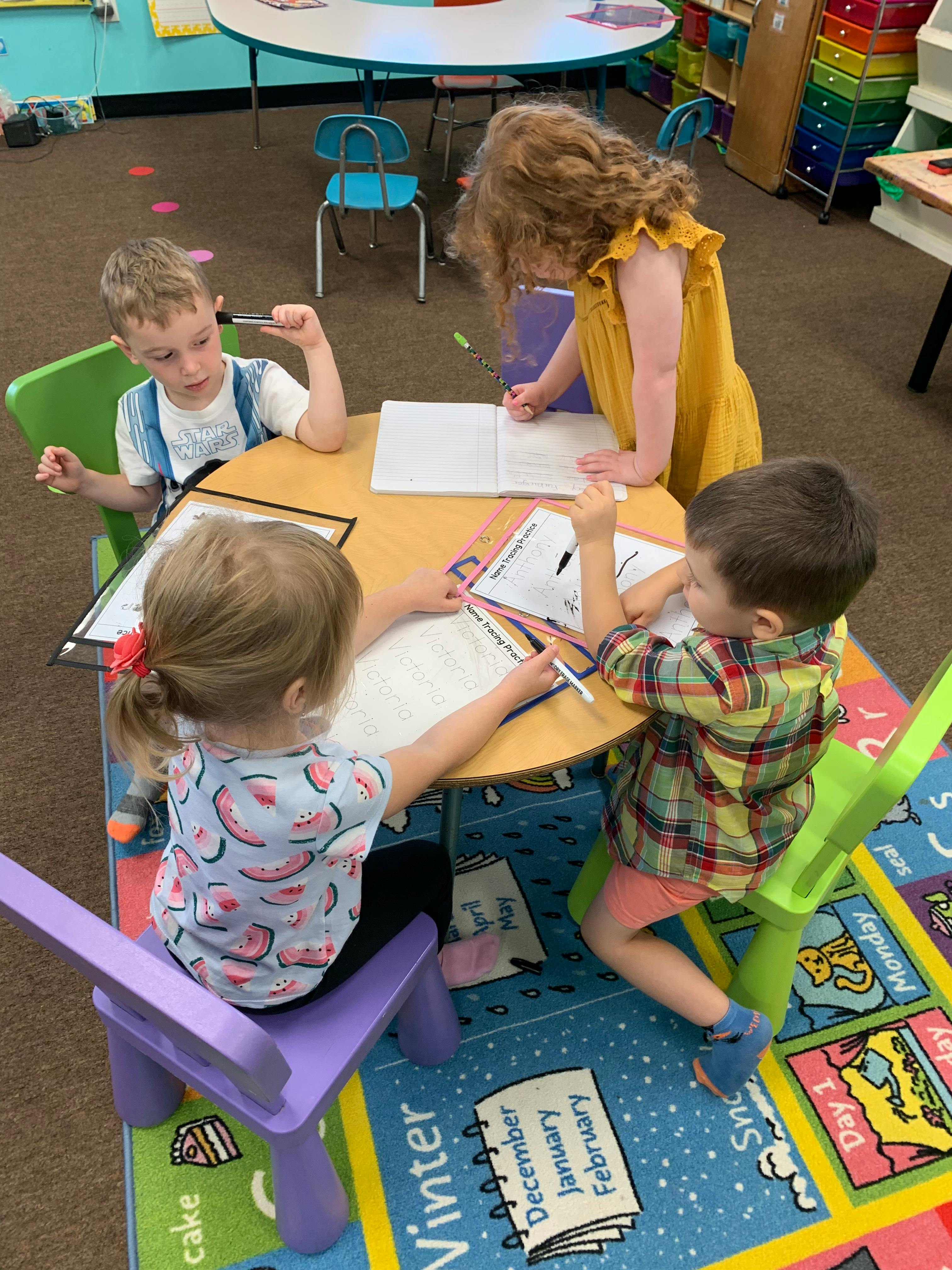 Four children drawing and writing at a small round table in a classroom