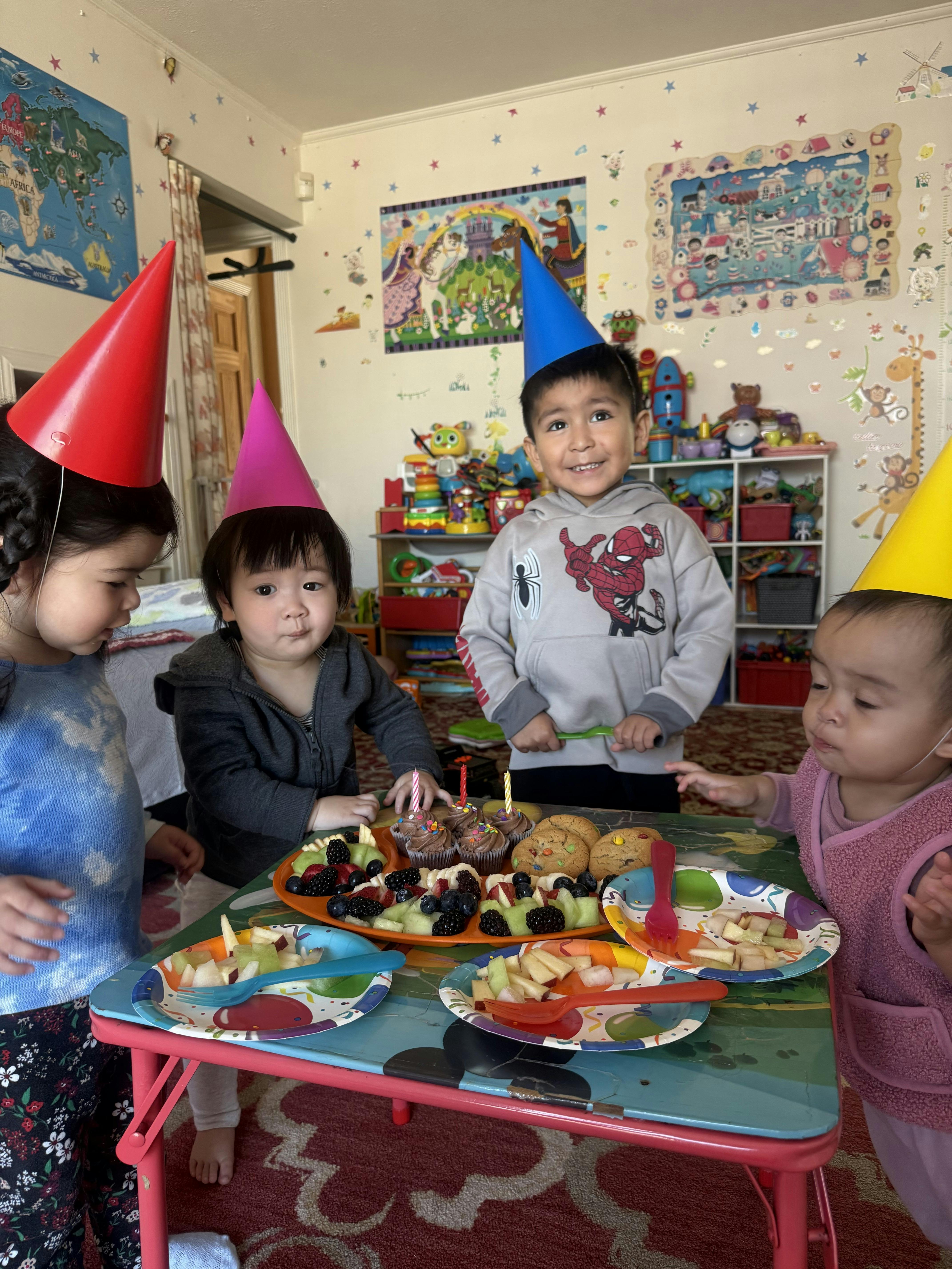 Four children in party hats gathered around a table with fruit, cupcakes, and cookies