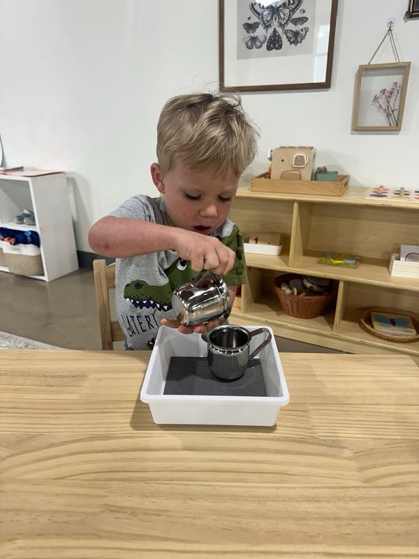 Child pouring from a small metal pitcher into a cup in a tray