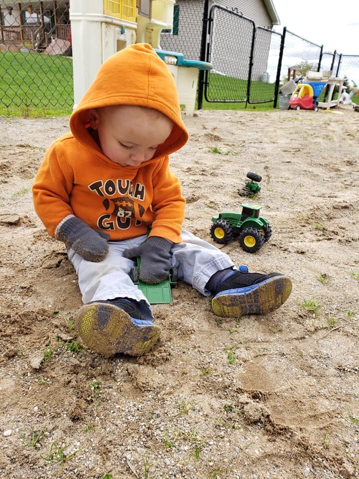 Child in orange hoodie and mittens sitting in dirt with toy tractor