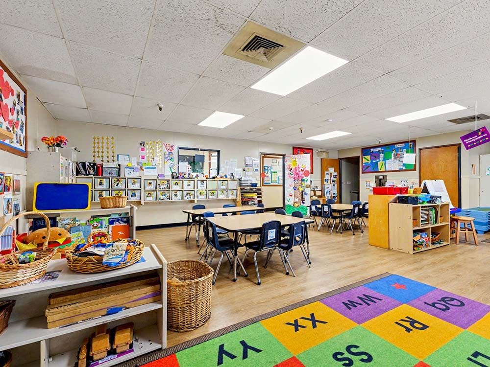 Classroom with small tables and chairs, shelves with toys, and an alphabet rug