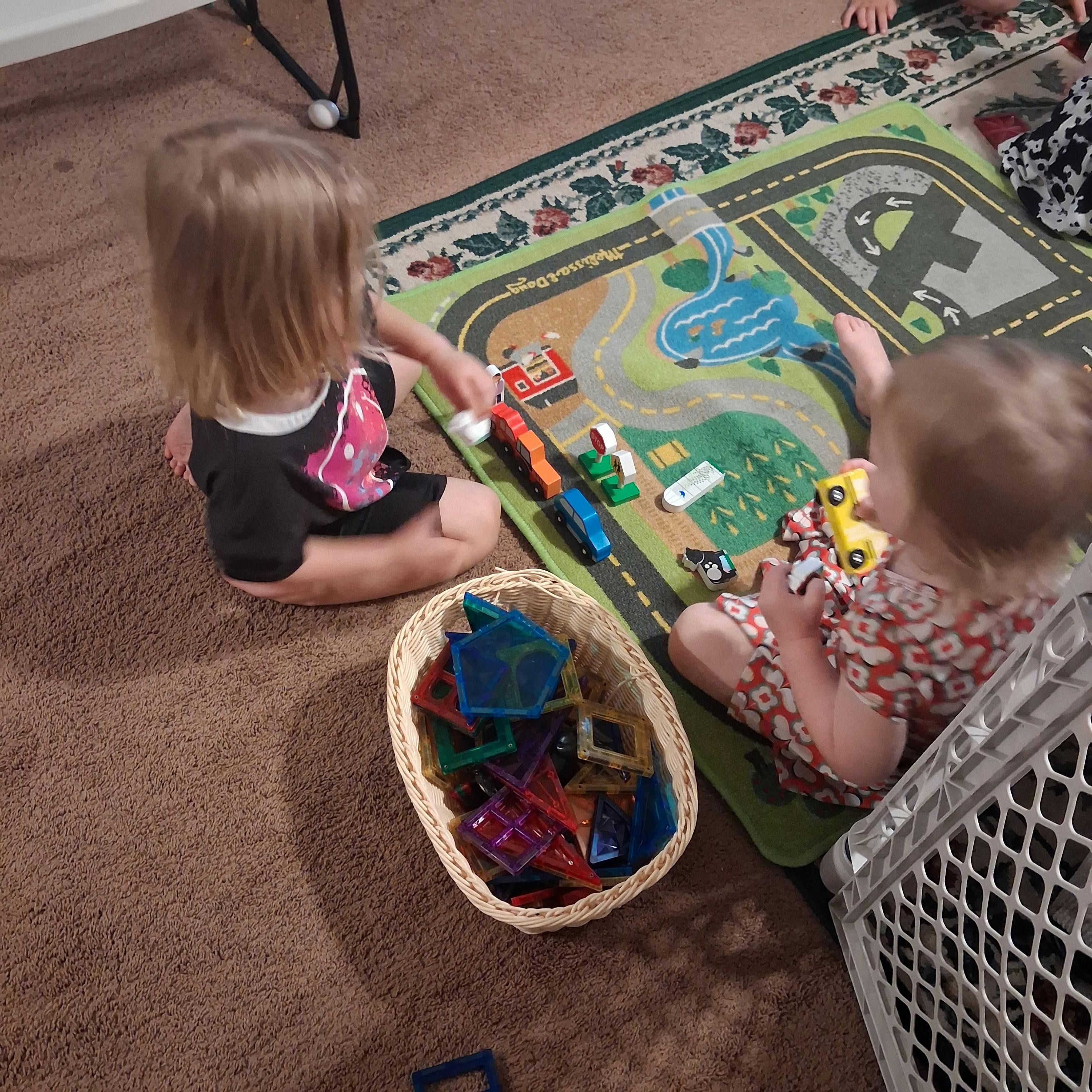 Two children playing with toy vehicles on a rug and magnetic tiles in a basket