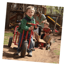 Child standing with a red toy vehicle, another child and adult in background