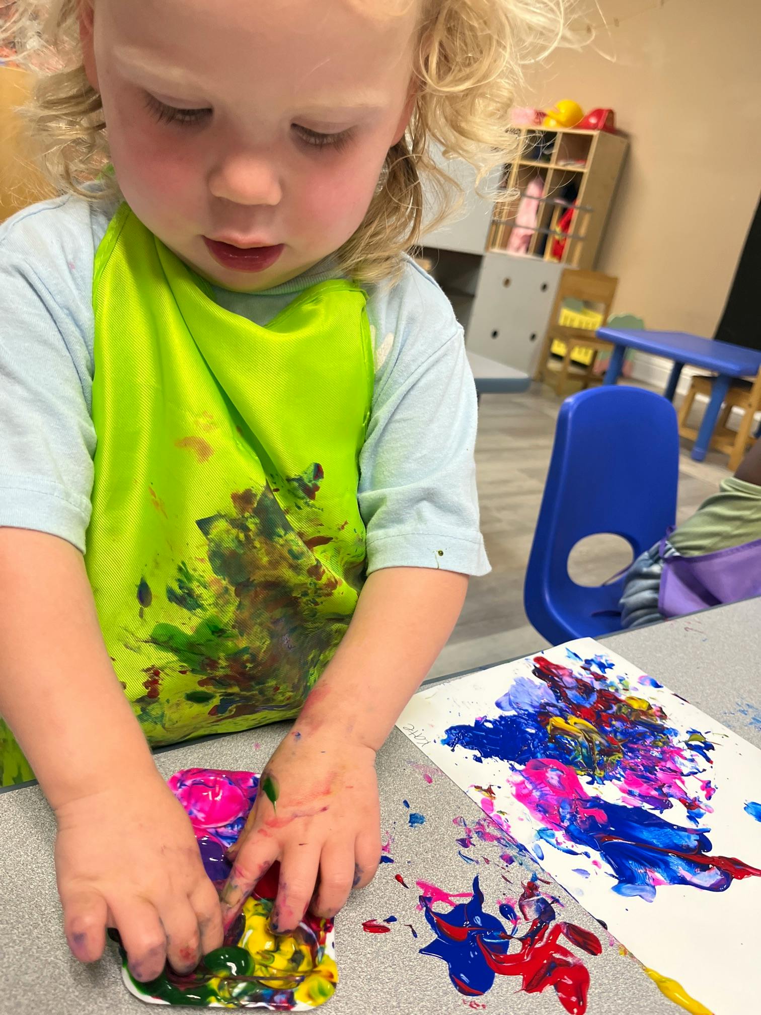 Child wearing an apron finger painting at a table in a classroom