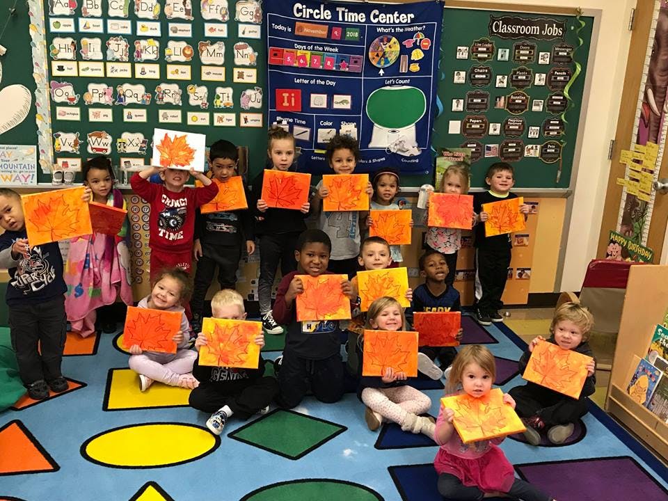 Many children in a classroom holding up orange and yellow artwork