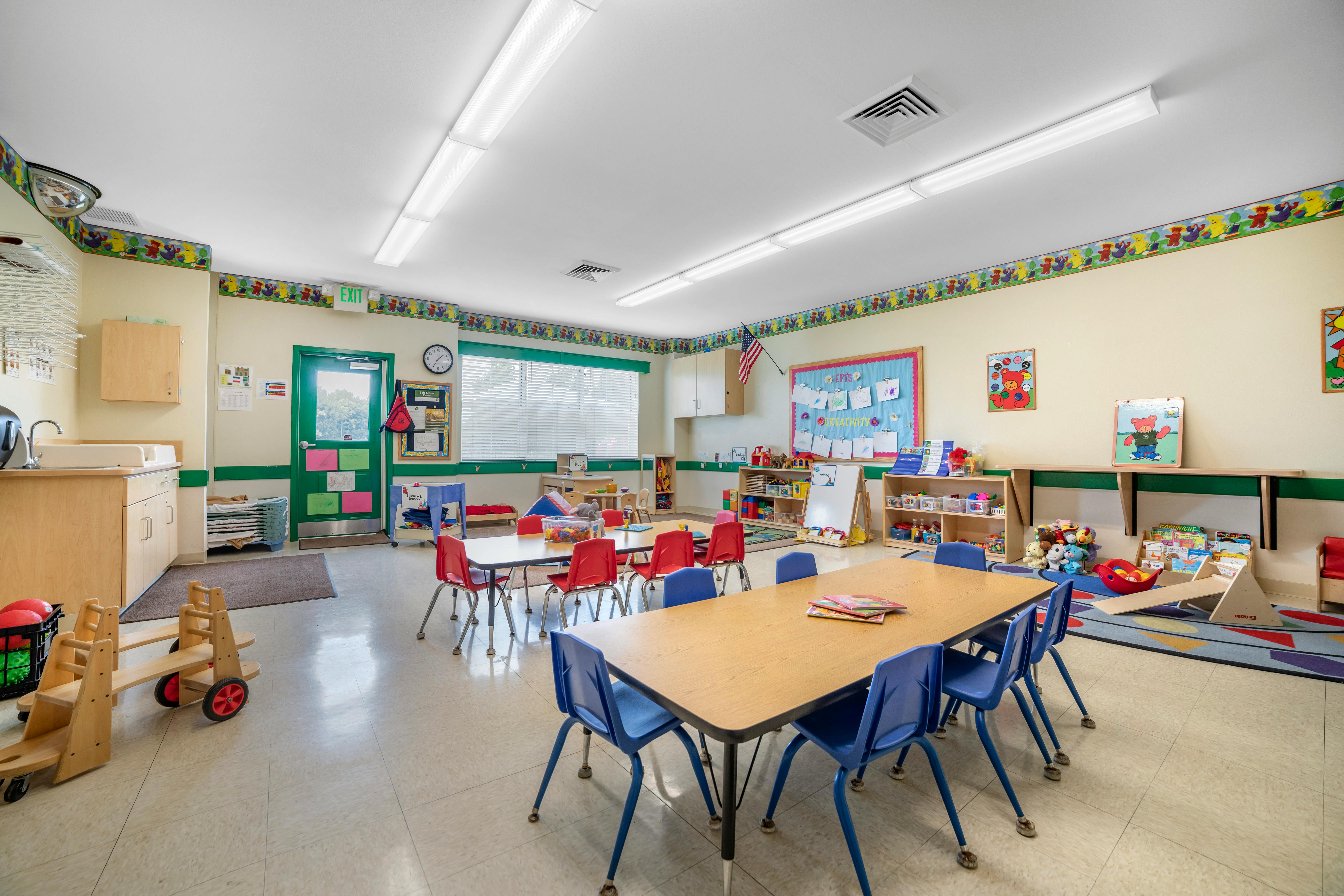 A colorful empty classroom with small tables, chairs, and shelves full of toys and books